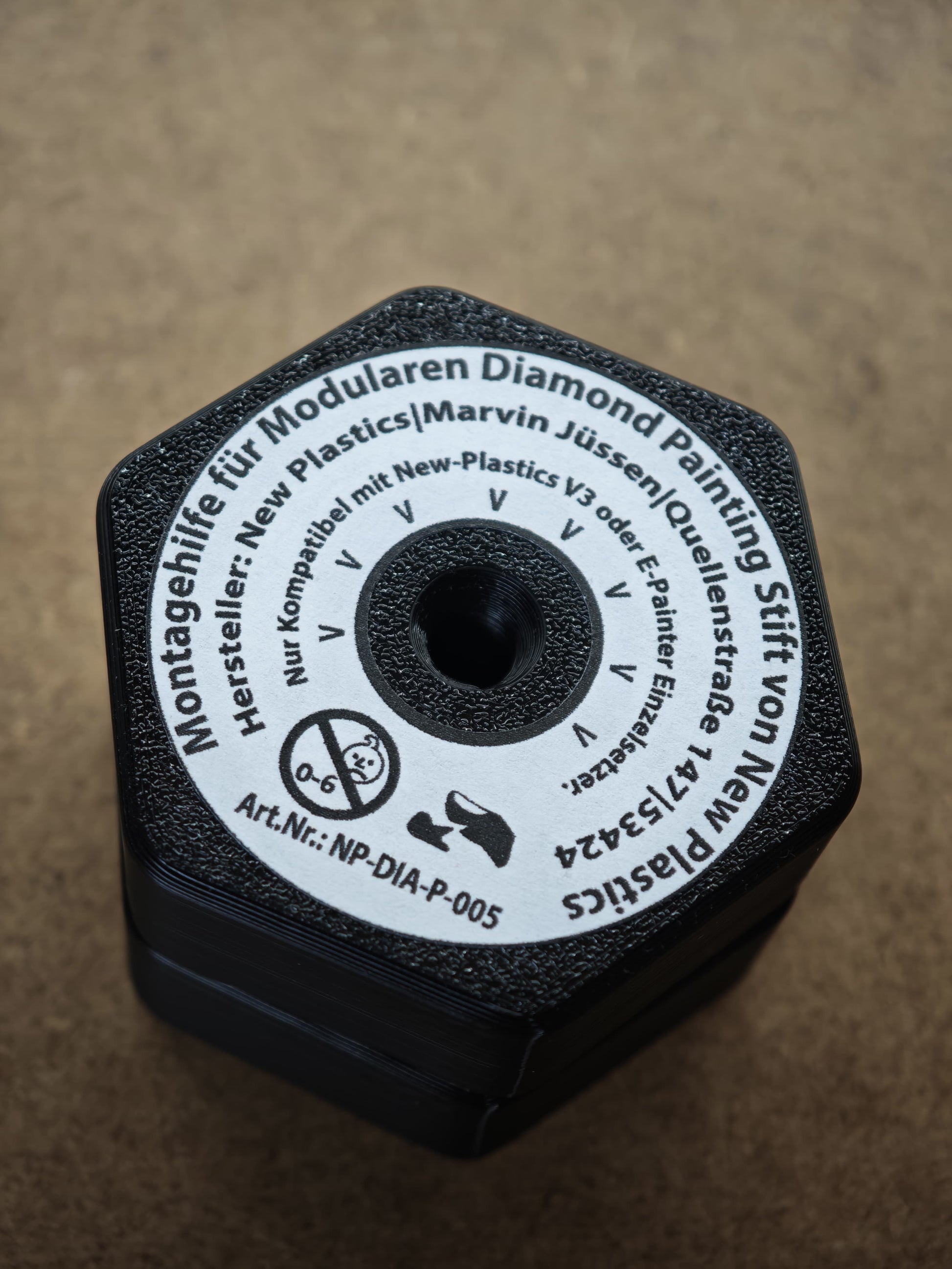 Eine Nahaufnahme zeigt eine sechseckige schwarze Montagehilfe für Modularen Diamond Painting Stift von New Plastics aus Kunststoff, die auf Deutsch mit Marken- und Produktdetails beschriftet ist. Das Montagewerkzeug von New Plastics liegt auf einer braunen Oberfläche.