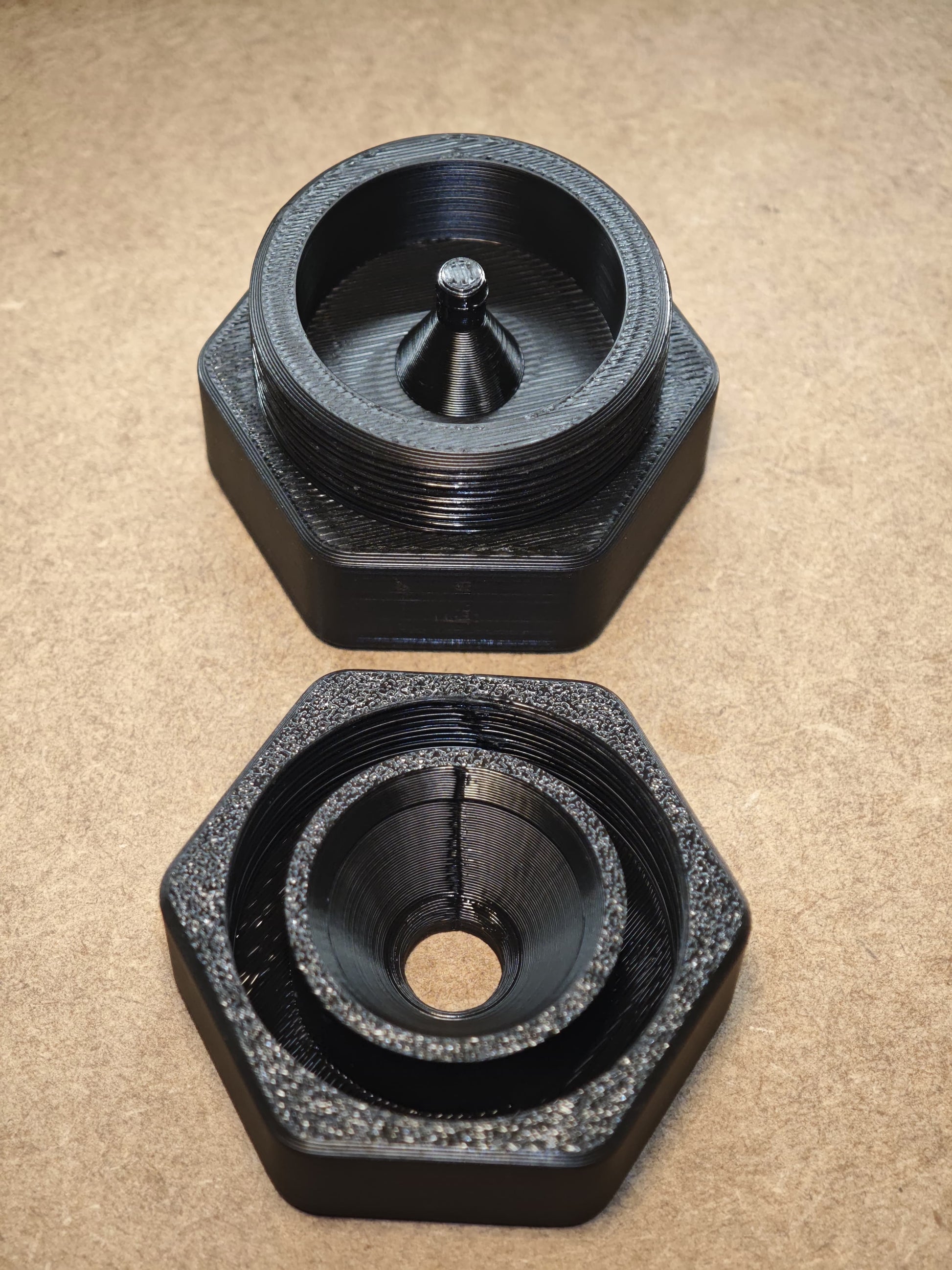 Abgebildet sind zwei schwarze, sechseckige, 3D-gedruckte Montagehilfen für den modularen Diamond Painting Stift von New Plastics. Eine verfügt über einen zentralen Kegel, die andere über ein Loch, was eine nahtlose Montage ermöglicht. Beide haben Gewindekanten und liegen auf einer braunen Oberfläche.