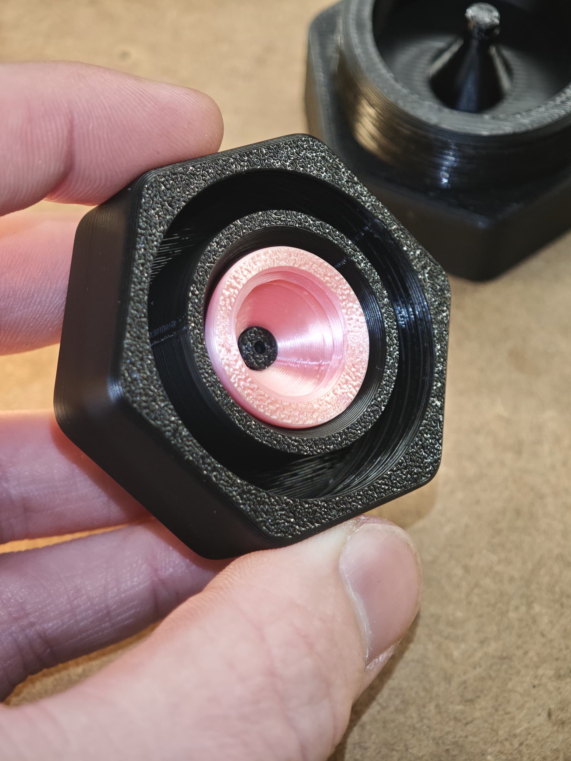 Eine Nahaufnahme einer Hand, die eine schwarze sechseckige „Montagehilfe für Modularen Diamond Painting Stift von New Plastics“ mit einer rosa-metallischen kegelförmigen Mitte von New Plastics hält; ein weiteres ähnliches Werkzeug ist im Hintergrund auf einer braunen Oberfläche unscharf zu sehen.