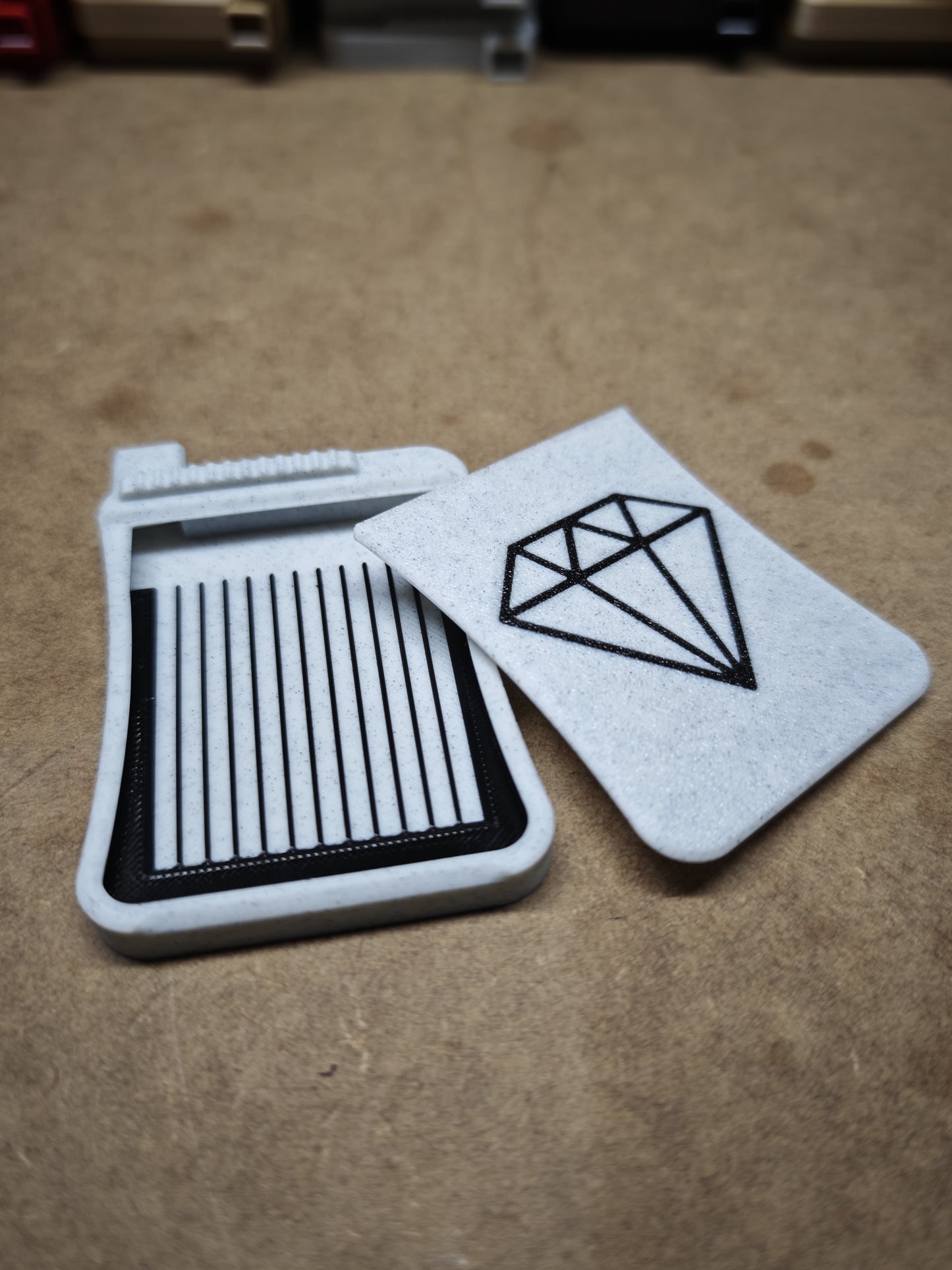 Auf einer braunen Oberfläche liegen zwei Untersetzer aus weißem Filz; einer mit einer schwarzen Rautenkontur, die die Präzision des Diamond Painting Schiffchen S3 von New Plastics widerspiegelt, und der andere mit schwarzen vertikalen Linien wie Notizbuchseiten.