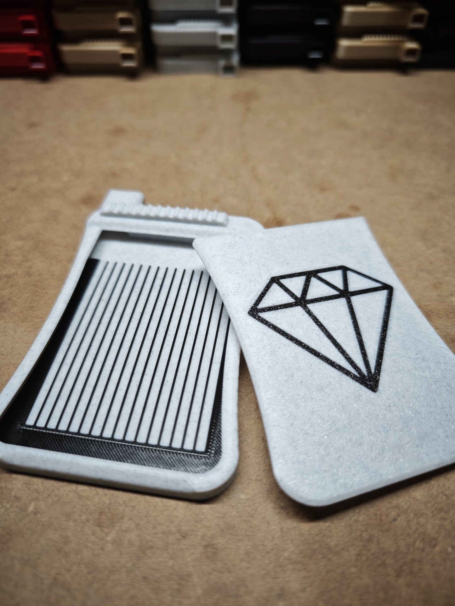 Zwei graue Behälter von New Plastics stehen auf einer Fläche. Einer davon ist das Diamond Painting Schiffchen S3 Plus mit einem großen schwarzen Rautenmuster. Der andere hat oben schmale, parallele Schlitze, vor dem Hintergrund unscharfer Regale mit verschiedenen Gegenständen.