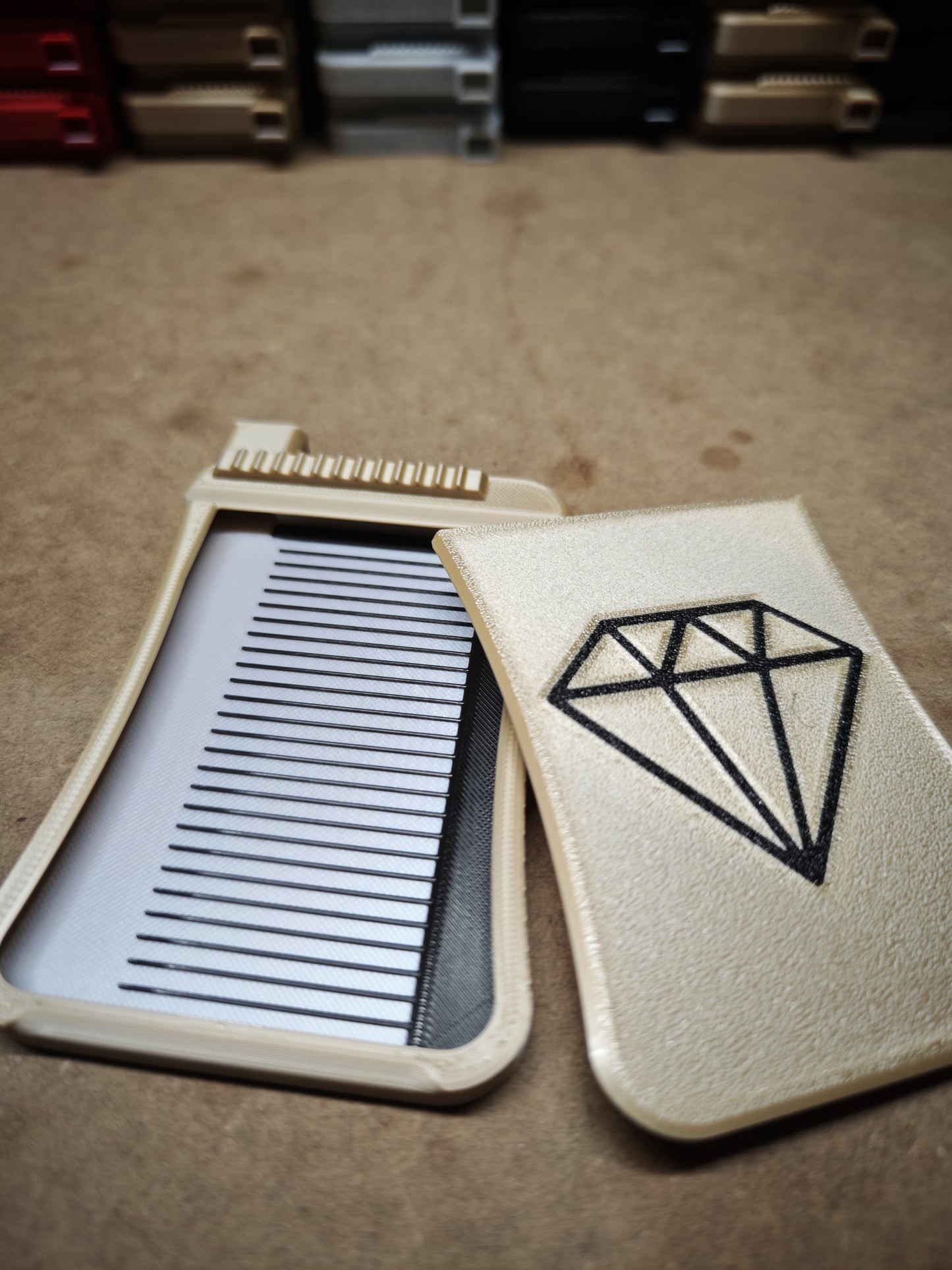 Ein beiges, offenes Gehäuse von New Plastics Präzision mit einem geometrischen Design ähnlich dem Diamond Painting Schiffchen S3 Plus hält einen eleganten schwarz-silbernen Kamm auf einem Tisch, während im Hintergrund mehrere andere Gehäuse gestapelt sind.