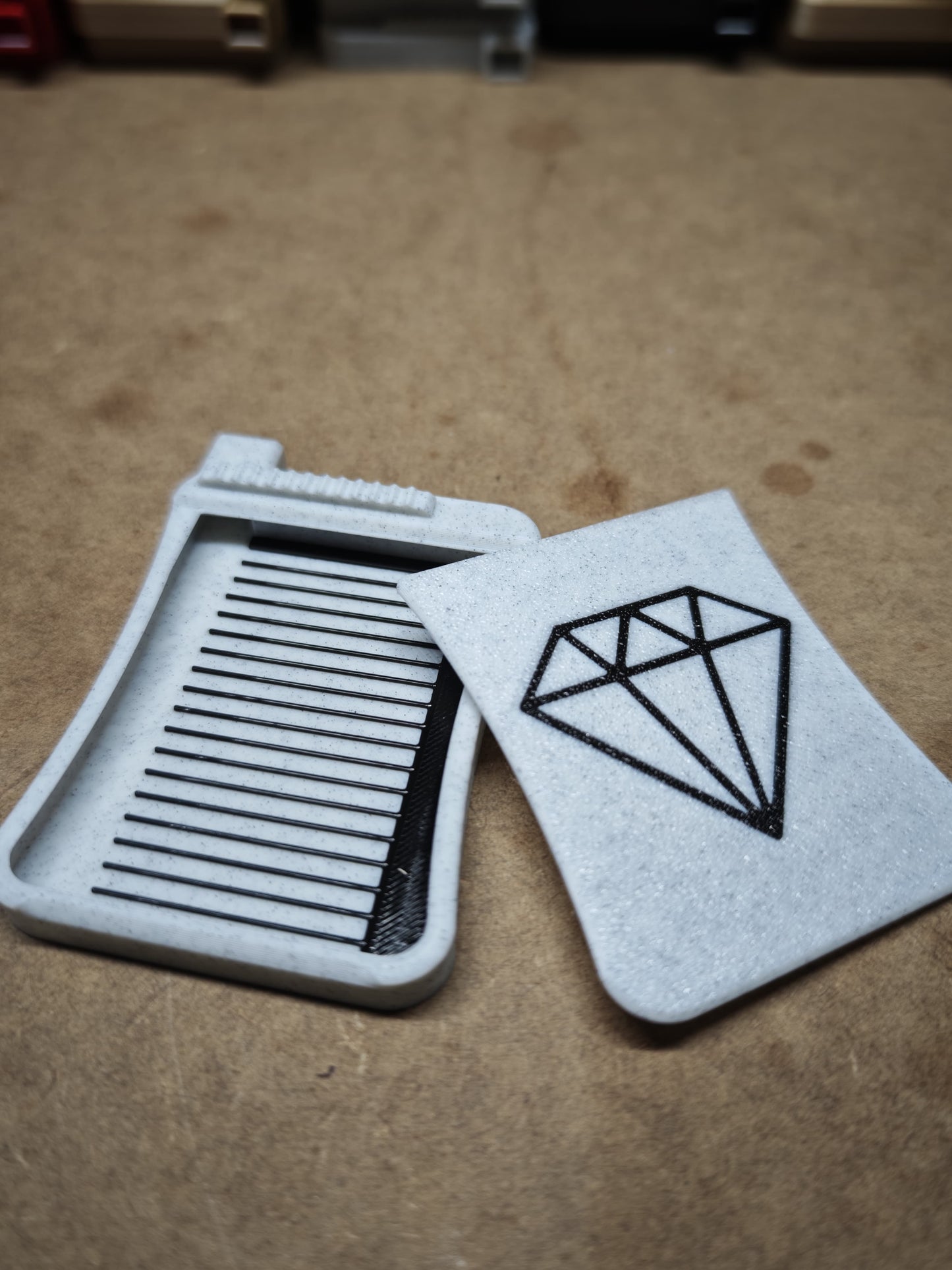 Zwei weiße Kunststoffobjekte liegen auf einer strukturierten Oberfläche: ein Kamm und ein New Plastics Diamond Painting Schiffchen S3, kartenförmig mit einem eleganten schwarzen geometrischen Rautenmuster.