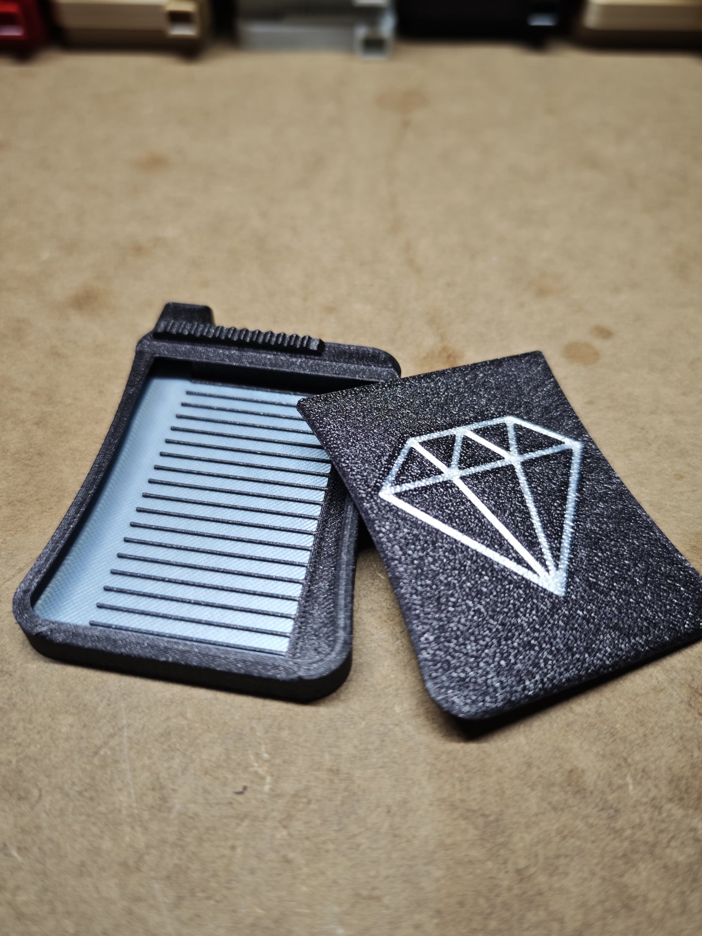 Zwei schwarze, rechteckige New Plastics-Objekte liegen auf einer braunen Oberfläche. Das untere Objekt ähnelt dem Diamond Painting Schiffchen S3 mit geriffelter Innenseite und besticht durch Präzision und Komfort. Das darüberliegende Objekt zeigt ein filigranes weißes Diamond Painting-Design, ideal für den Schiebe-Stopper für den Ausguss.