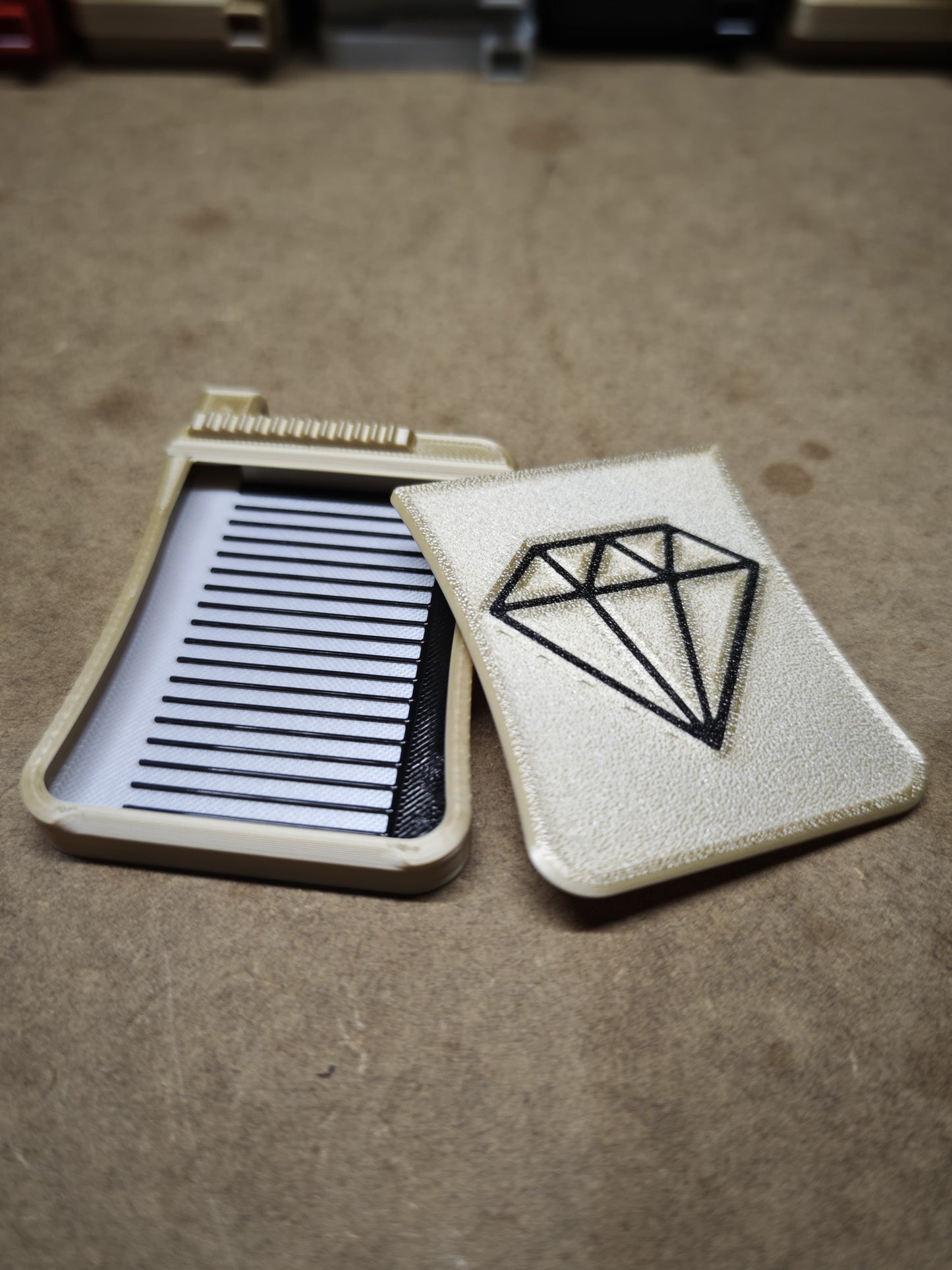 Ein weißes Kunststoffgehäuse von New Plastics mit einer Diamantillustration liegt auf einer braunen Oberfläche. Im Inneren des Diamond Painting Schiffchen S3-Gehäuses ist ein schwarzer Kamm für Präzision und Komfort teilweise sichtbar.