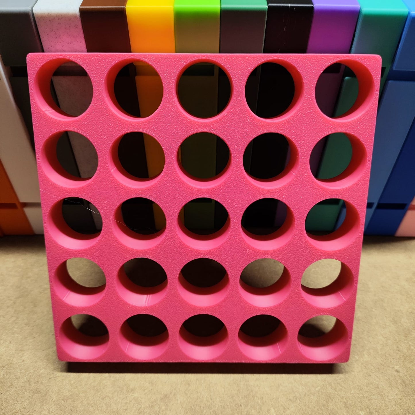 Die Steinchen-Garage Modulplatte von New Plastics ist ein rosa Kunststoffregal für 25 Farbdosen rund mit 6 Reihen und 4 Spalten mit runden Löchern. Es besteht aus leuchtenden gestapelten rechteckigen Blöcken in Orange, Grün und Lila – das perfekte Organisationssystem für jeden kreativen Raum.