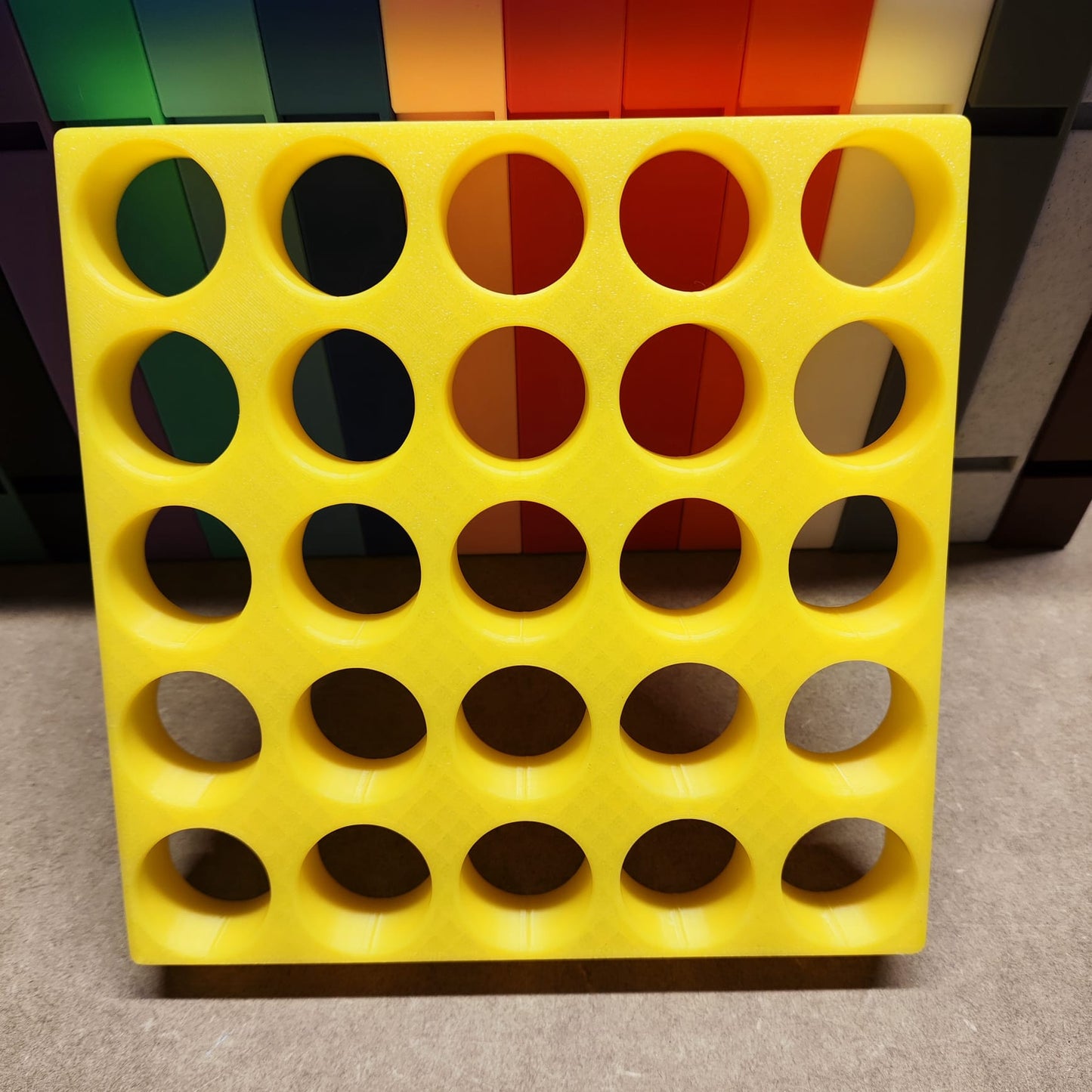 Die Steinchen-Garage Modulplatte für 25 Farbdosen Rund von New Plastics besteht aus einem gelben quadratischen Gitter mit runden Löchern über bunten vertikalen Streifen, die in einem 5x5-Muster in Grün-, Orange- und Rottönen angeordnet sind.