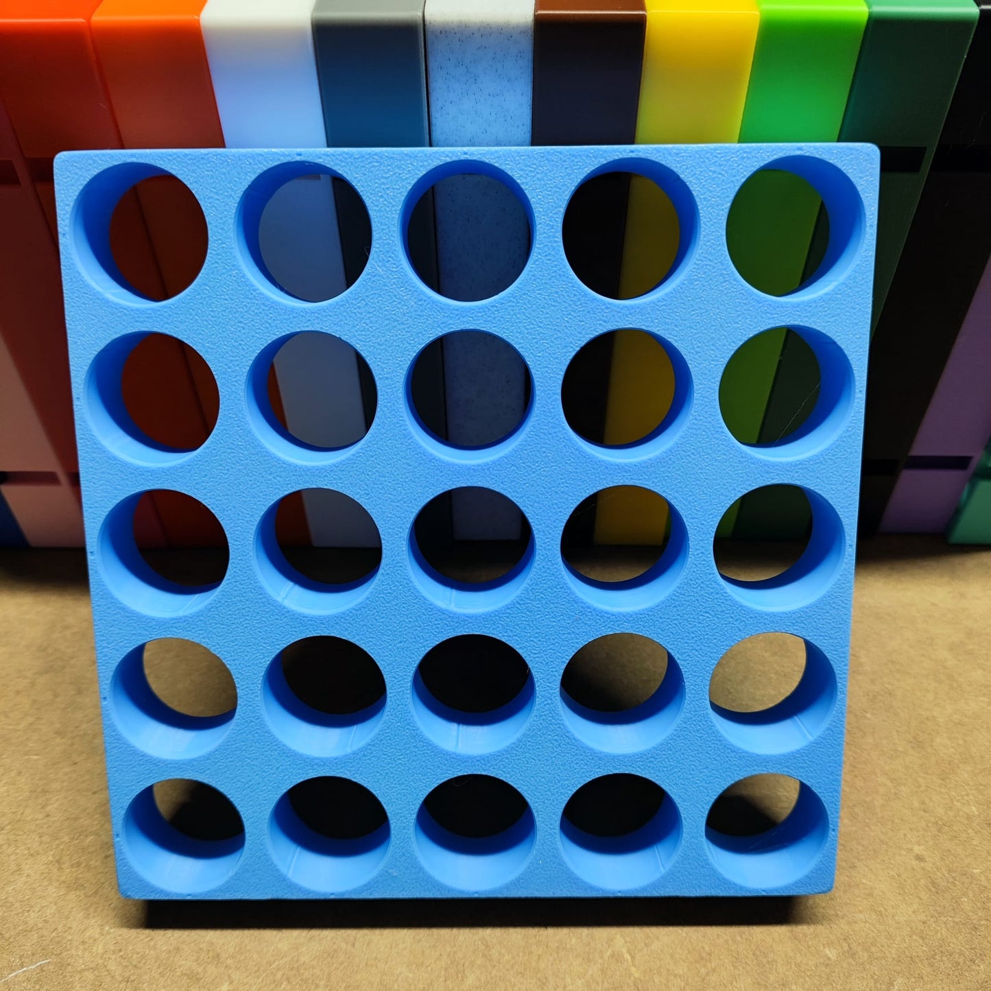 Eine Steinchen-Garage Modulplatte für 25 Farbdosen Rund von New Plastics liegt auf einer braunen Oberfläche und zeigt ein blaues Gitter mit kreisförmigen Löchern. Bunte rechteckige Blöcke im Hintergrund bilden ein geordnetes Muster, das an Diamantmalerei erinnert.