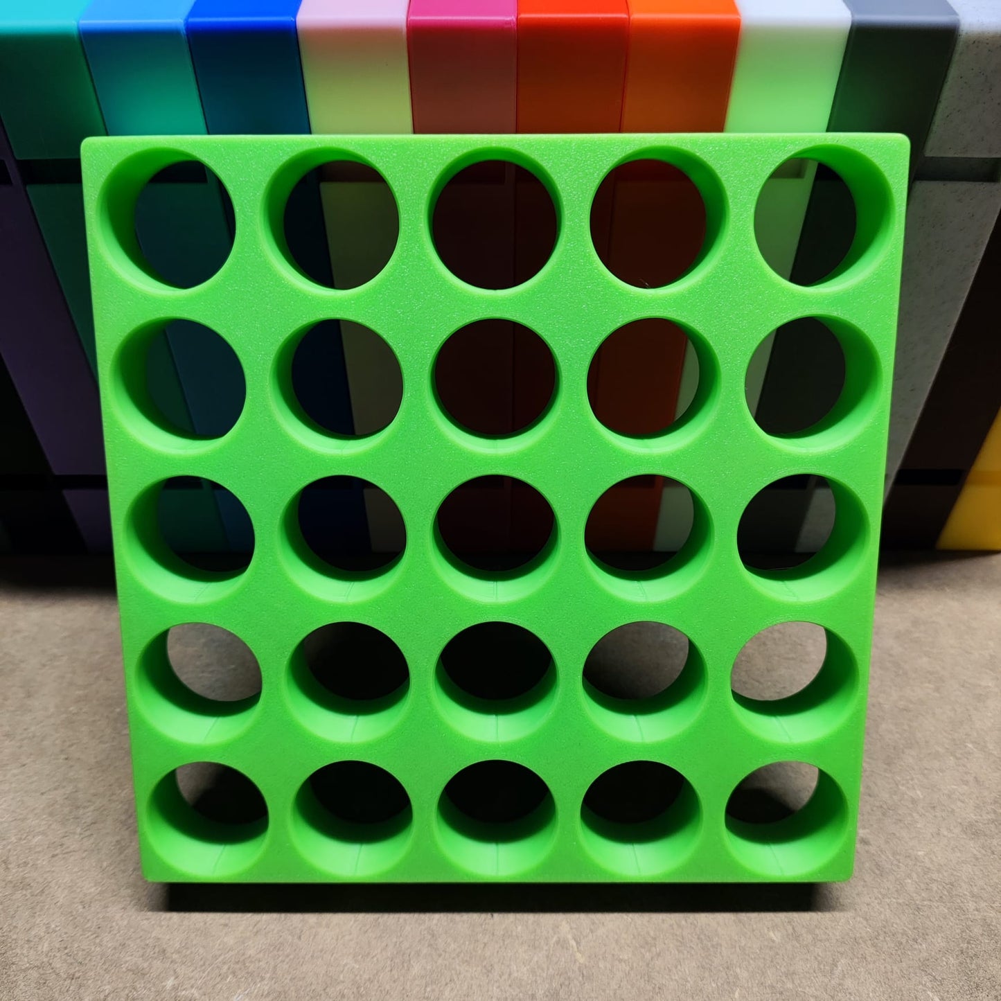 Die Steinchen-Garage Modulplatte von New Plastics ist ein grünes Kunststoffgitter mit kreisrunden Löchern, die vor einem Hintergrund aus bunten rechteckigen Blöcken angeordnet sind und ähnlich einem Diamantgemälde blaue, rote und orange Farbtöne auf Beige aufweisen.