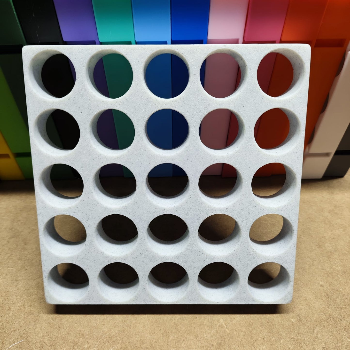 Eine Steinchen-Garage Modulplatte für 25 Farbdosen Rund von New Plastics, die an ein effizientes Organisationssystem erinnert, ruht auf einer Holzoberfläche. Dahinter sind Reihen von Ordnern in Blau, Grün, Rot, Rosa und Schwarz zu sehen.