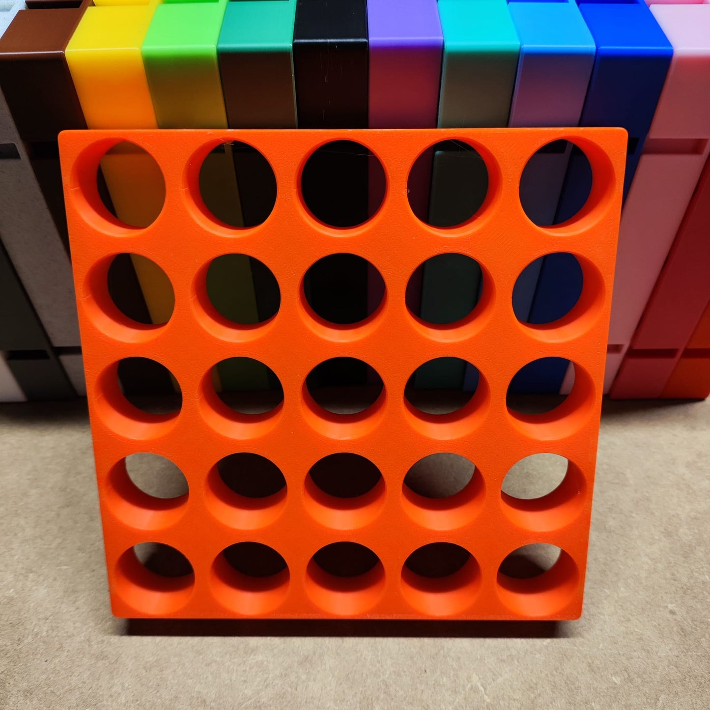 Eine leuchtend orangefarbene New Plastics Steinchen-Garage Modulplatte für 25 Farbdosen Rund mit einem Raster aus 25 kreisförmigen Löchern sticht im Vordergrund hervor. Dahinter befindet sich ein auffälliger Stapel bunter rechteckiger Blöcke in den Farbtönen Gelb, Grün, Schwarz, Blau und Rosa.