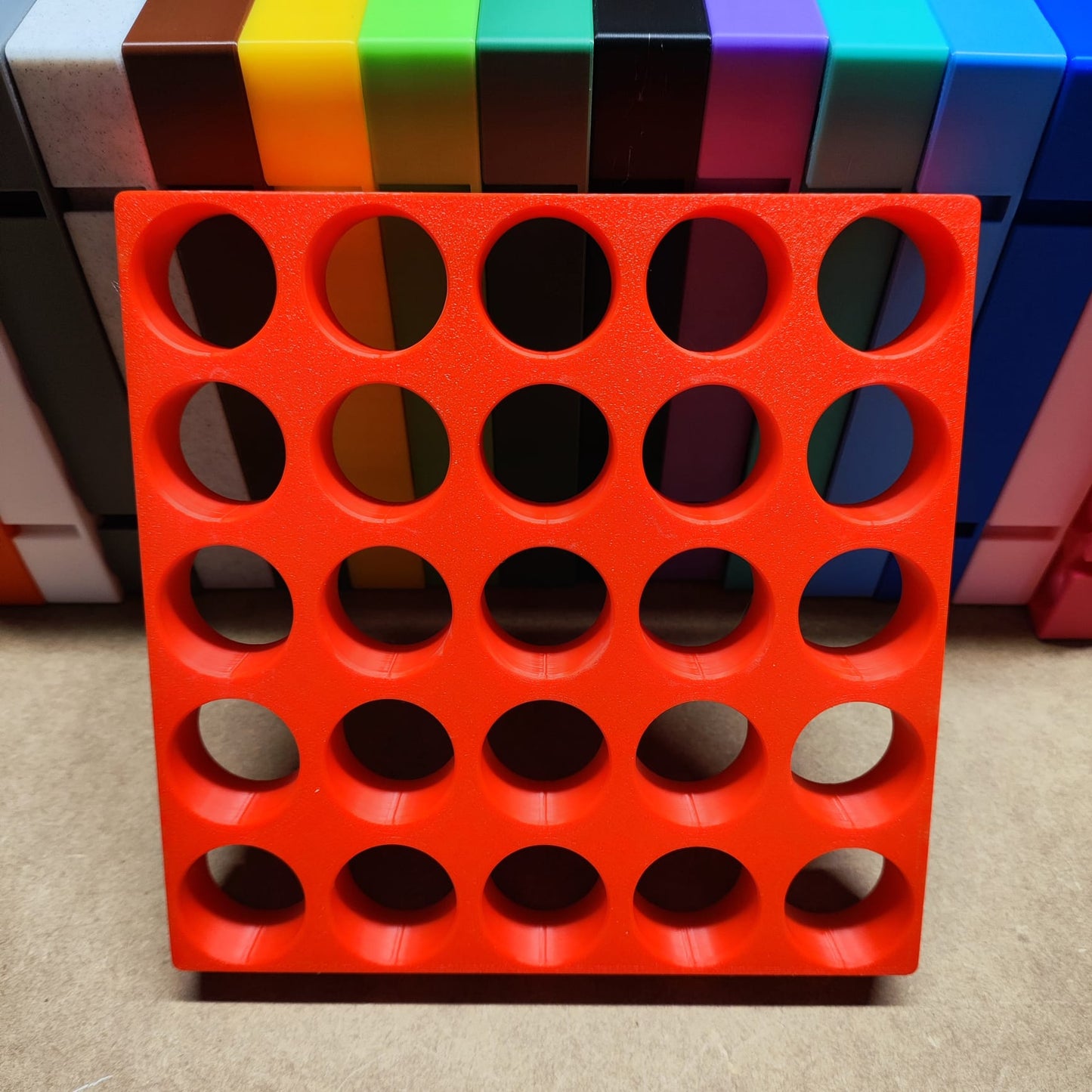 Vor einem bunten Blockhintergrund wird eine Steinchen-Garage Modulplatte für 25 Farbdosen Rund von New Plastics angezeigt, die kreisförmige Löcher in einem roten Kunststoffquadrat wie ein Organisationssystem aufweist.