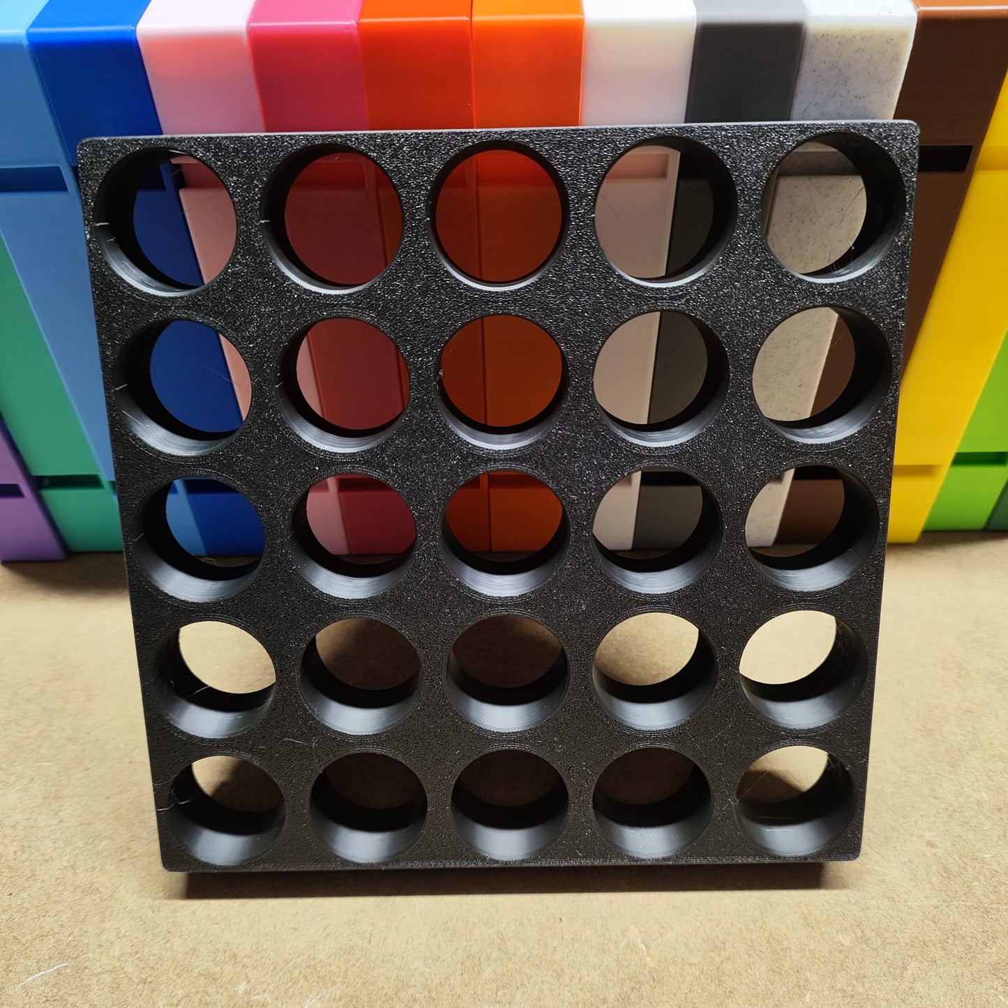 Die Steinchen-Garage Modulplatte für 25 Farbdosen Rund von New Plastics, ein schwarzes Kunststoffgitter mit kreisrunden Löchern, liegt als effektvolles Organisationssystem auf einer Fläche, im Hintergrund sind bunte rechteckige Platten vertikal angeordnet.