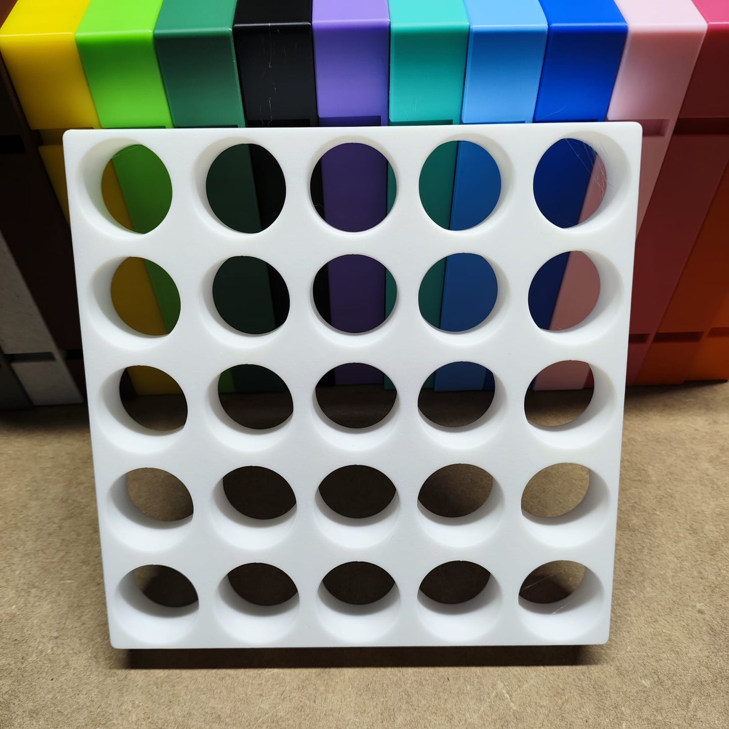 Die Steinchen-Garage Modulplatte für 25 Farbdosen Rund von New Plastics ist ein weißer Organizer mit runden Löchern, der aufrecht neben farbenfrohen, gestapelten rechteckigen Behältern steht. Der Hintergrund strahlt in Grün, Schwarz, Lila, Blau und Orange auf Beige – ideal für Bastel- oder Diamantmalerei-Fans.