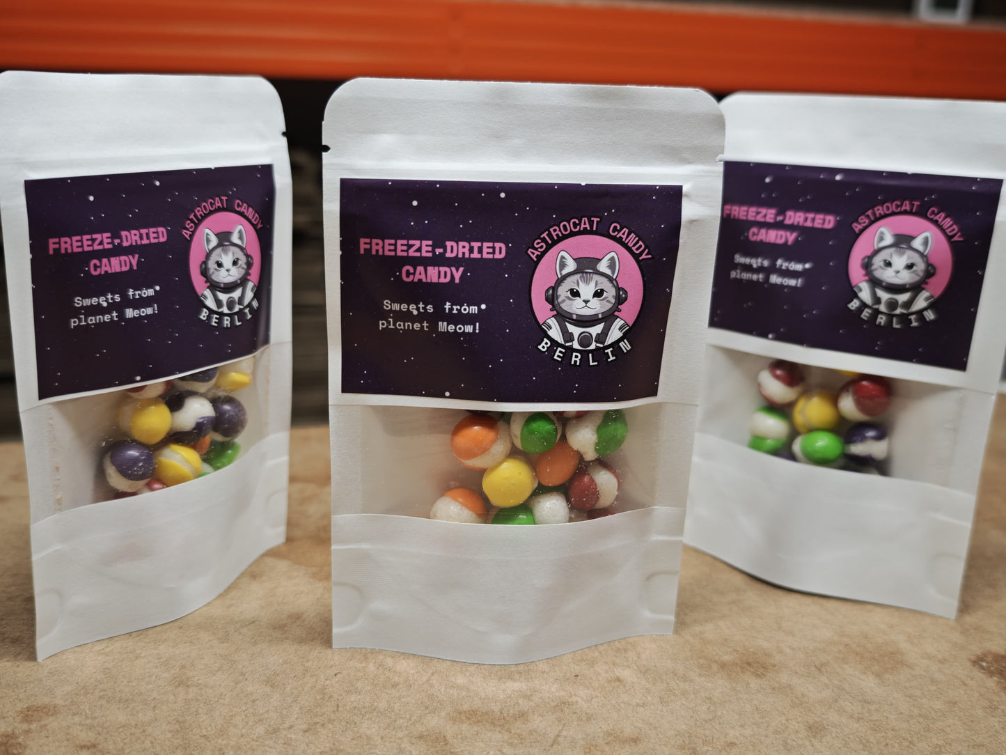 Drei 15-g-Beutel veganer Astrocat Candy Rainbow Bites präsentieren bunte Süßigkeiten durch transparente Fenster. Jeder Beutel zeigt eine Katze mit Astronautenhelm, den Text „Astrocat Candy Berlin“ und „Sweets from planet Meow!“ auf einem sternförmigen Fruchtgeschmack-Hintergrund.