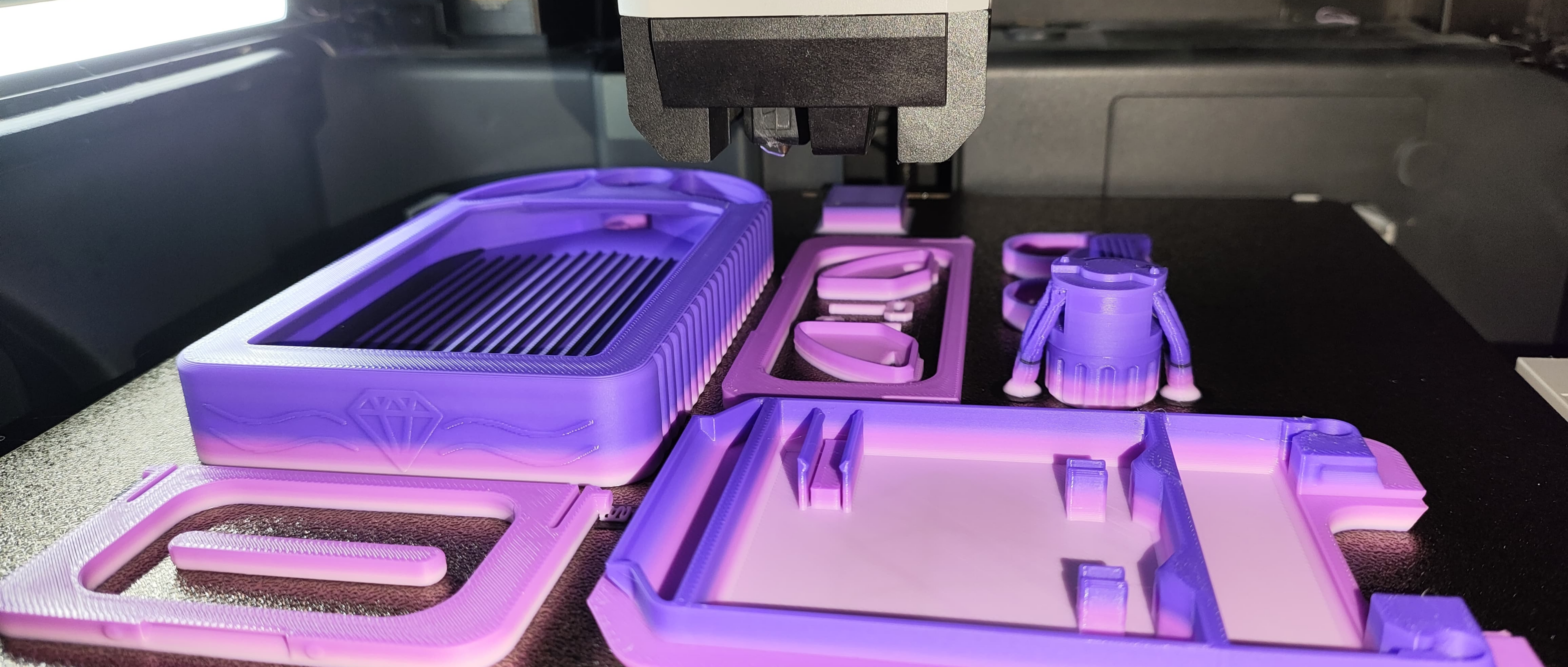 Ein 3D-Drucker fertigt auf einem schwarzen Druckbett mehrere violette Objekte, darunter einen Behälter und verschiedene Kleinteile. Die Objekte scheinen sich in unterschiedlichen Fertigstellungsstadien zu befinden, und ein helles Licht erhellt die Szene.