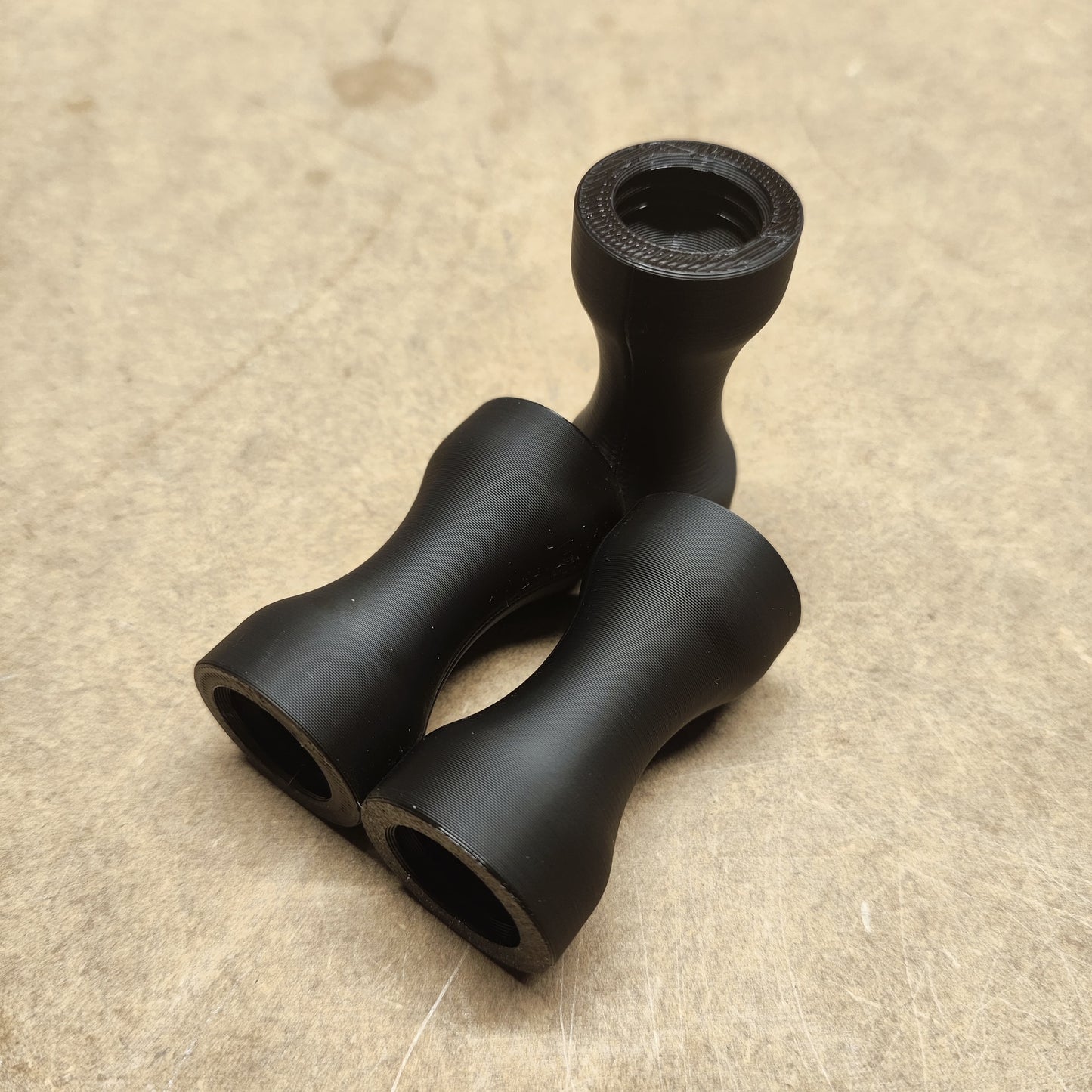 Drei Schmales Griffstück für Modularen Diamond Painting Stift von New Plastics, schwarz und zylindrisch mit ergonomischer Form, liegen auf einer hellen Holzoberfläche. Sie sehen identisch aus und sind in einer dreieckigen Formation angeordnet.