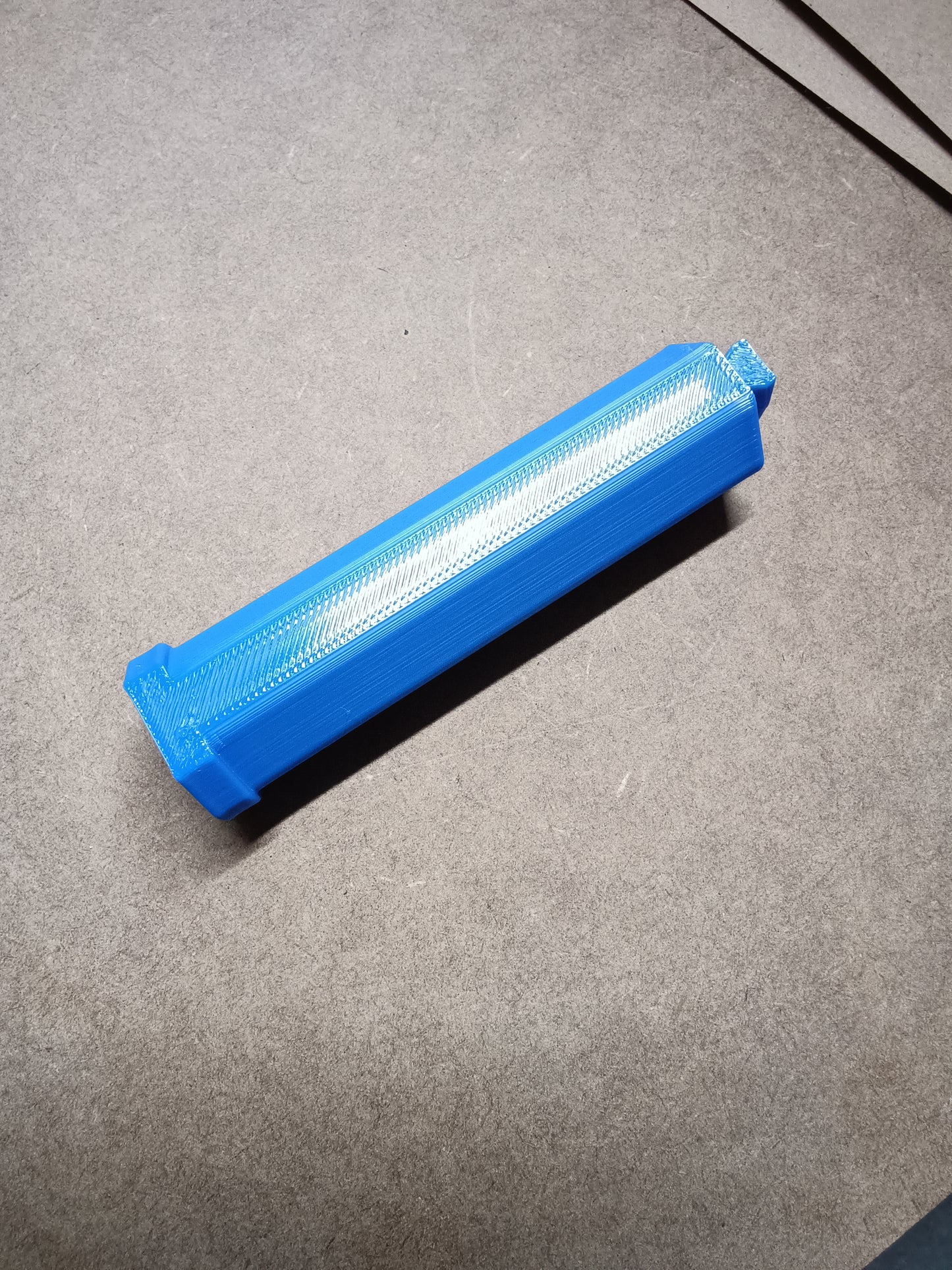 Ein langes, blaues 3D-gedrucktes Objekt, das einer Washi-Tape-Halterung für Steinchen Garage von New Plastics ähnelt, liegt auf einer grauen Oberfläche und zeigt seine flache rechteckige Form und komplizierte Textur.