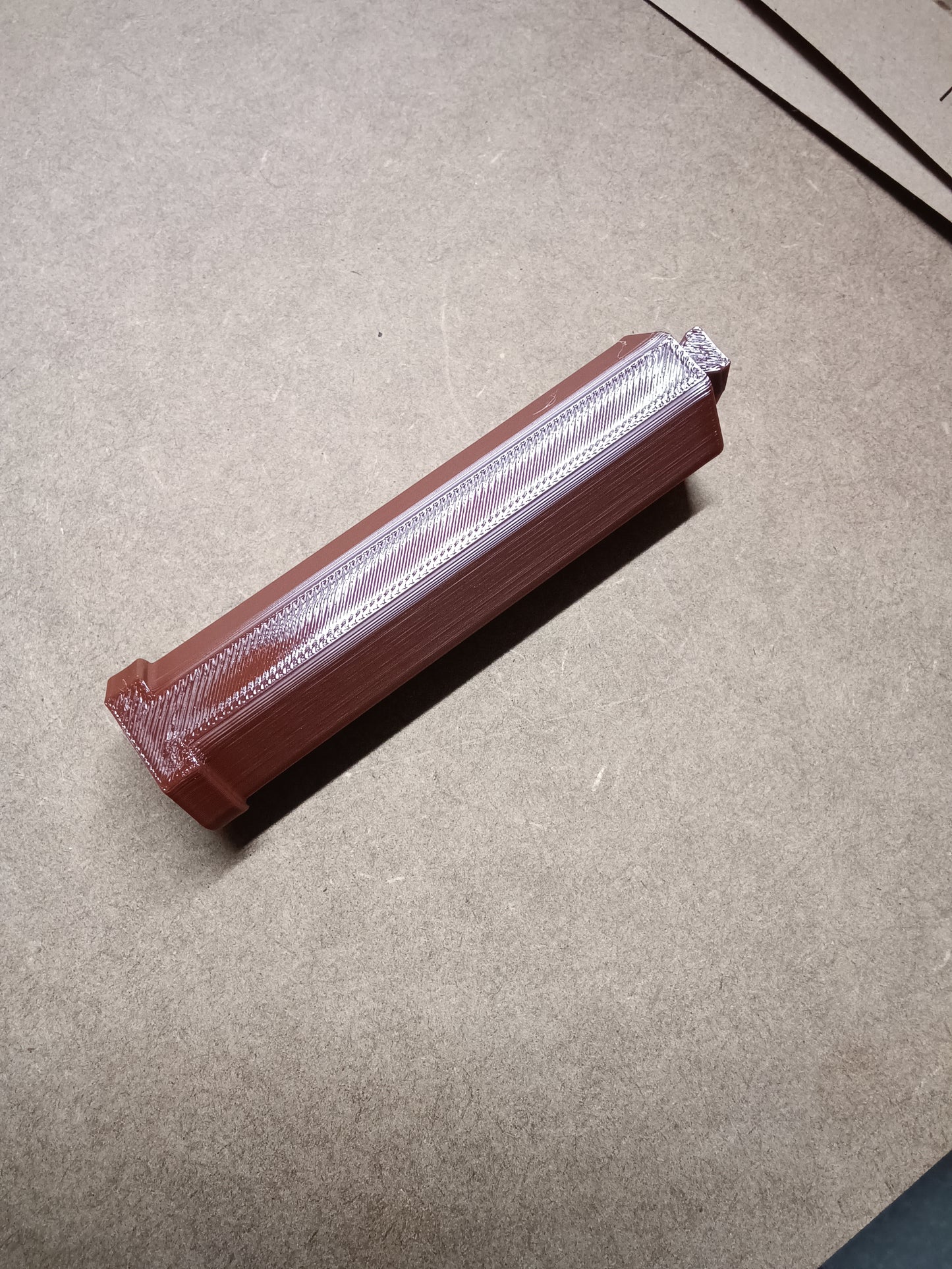 Ein brauner rechteckiger Artikel mit der Marke „Washi Tape Halterung für Steinchen Garage“ von New Plastics weist über die gesamte Länge einen transparenten Streifen auf und sitzt auf einer beigen Oberfläche mit einem strukturierten Muster auf der Oberseite.