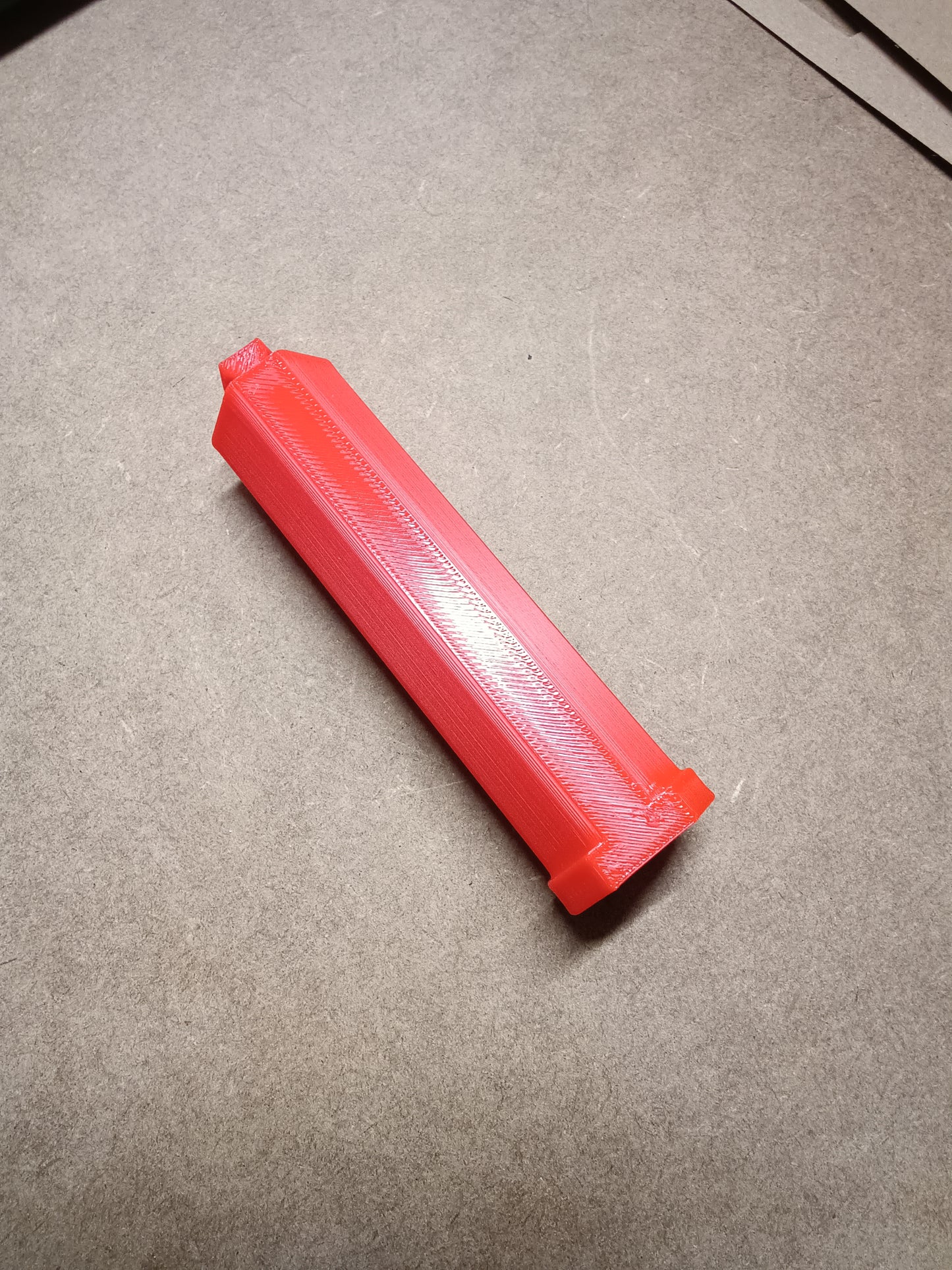 Eine rote, rechteckige 3D-gedruckte Washi Tape Halterung für Steinchen Garage von New Plastics mit strukturierter Oberfläche liegt auf einem braunen Hintergrund. Das Objekt ist länglich und hat an einem Ende eine spitze Spitze und am anderen eine flache Basis.