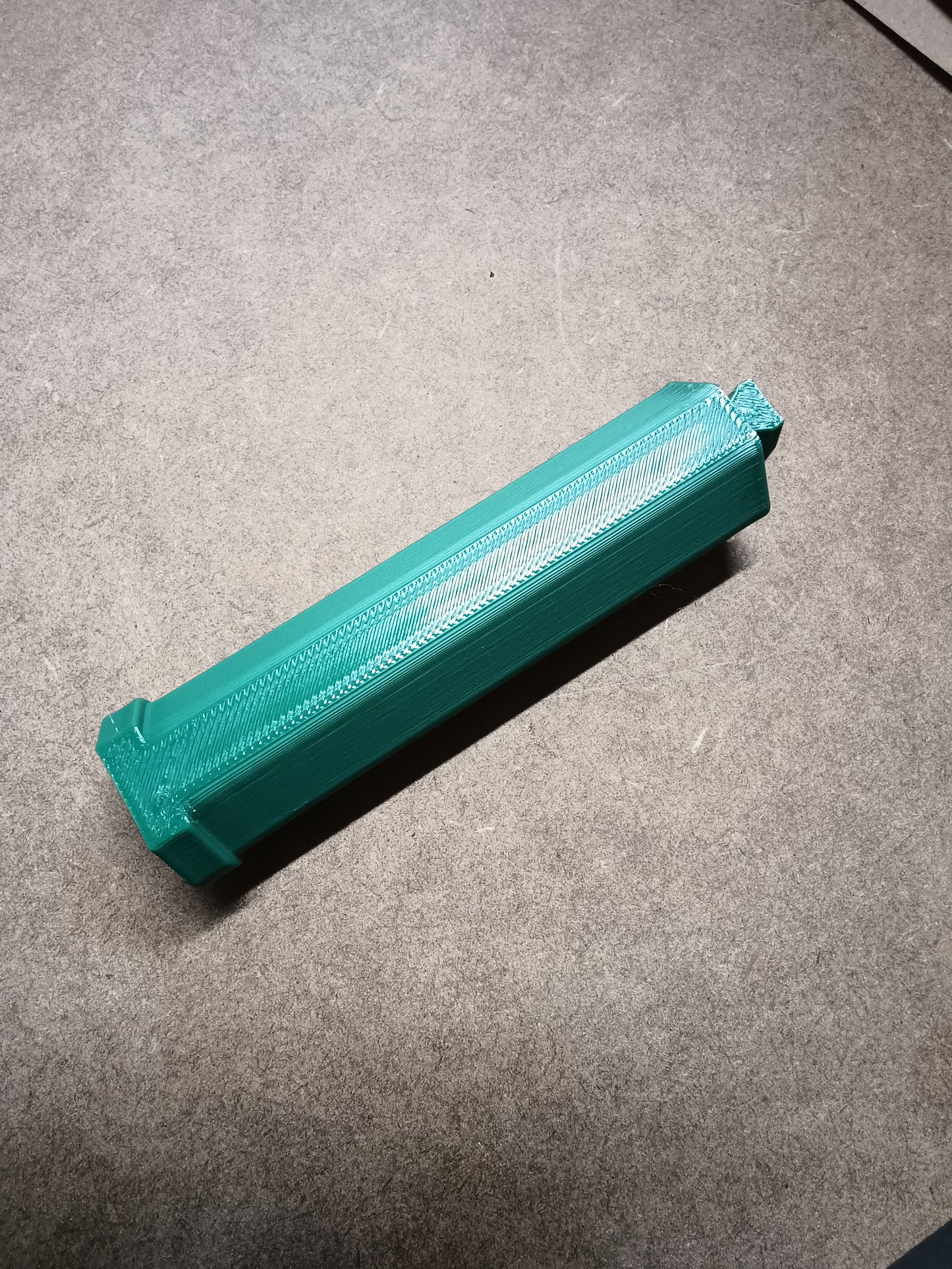 Eine Washi Tape Halterung für Steinchen Garage von New Plastics, ein grünes, 3D-gedrucktes, längliches sechseckiges Prisma, ruht auf einer strukturierten grauen Oberfläche. Es weist entlang seiner Länge Rillen und flache Enden auf und bietet eine künstlerische Note, die an Diamantmalerei erinnert.