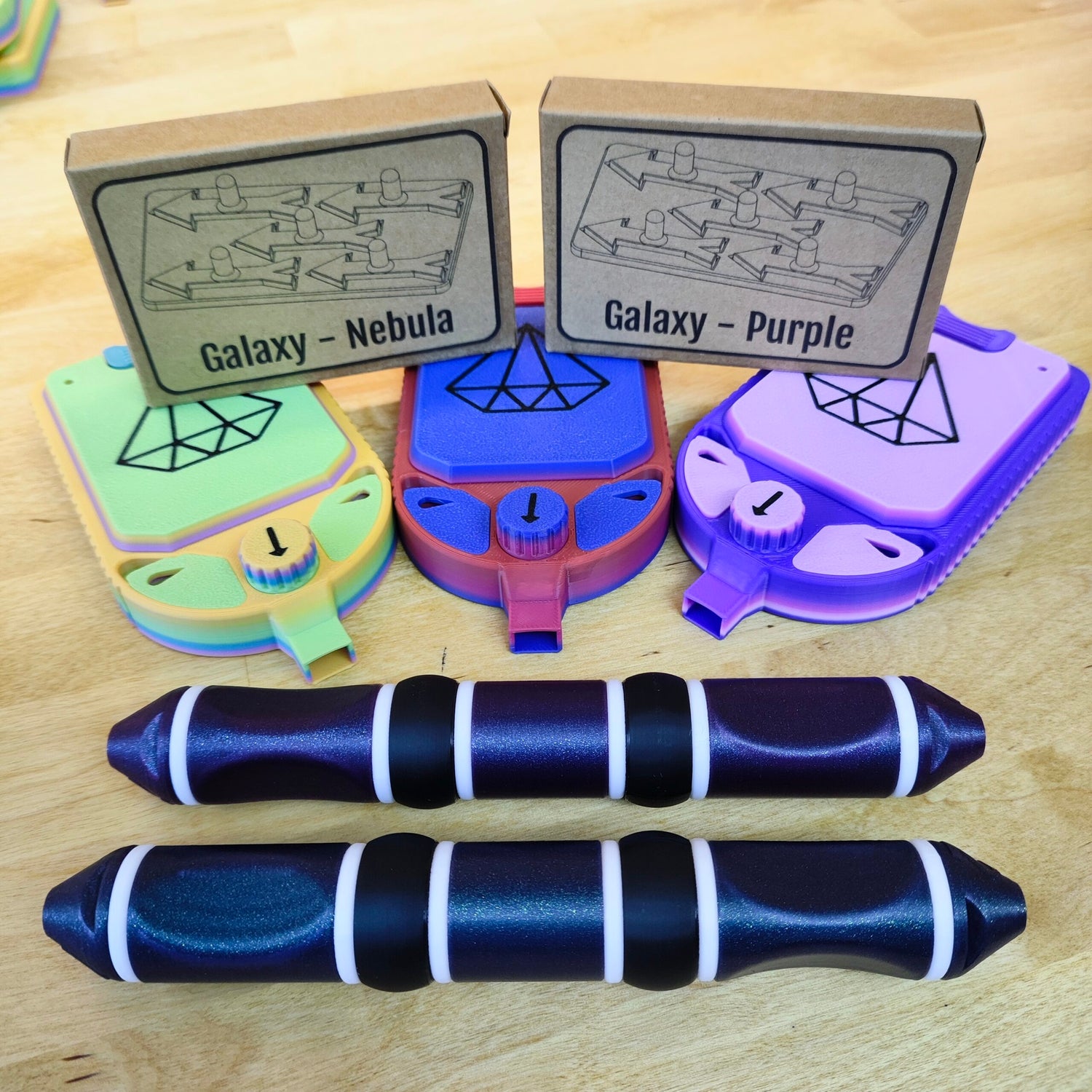 Bunte Handheld-Geräte mit geometrischen Mustern und Zifferblättern mit den Aufschriften „Galaxy Nebula“ und „Galaxy Purple“ werden auf einer Holzoberfläche präsentiert. Davor stehen zwei schwarze und violette zylindrische Objekte.