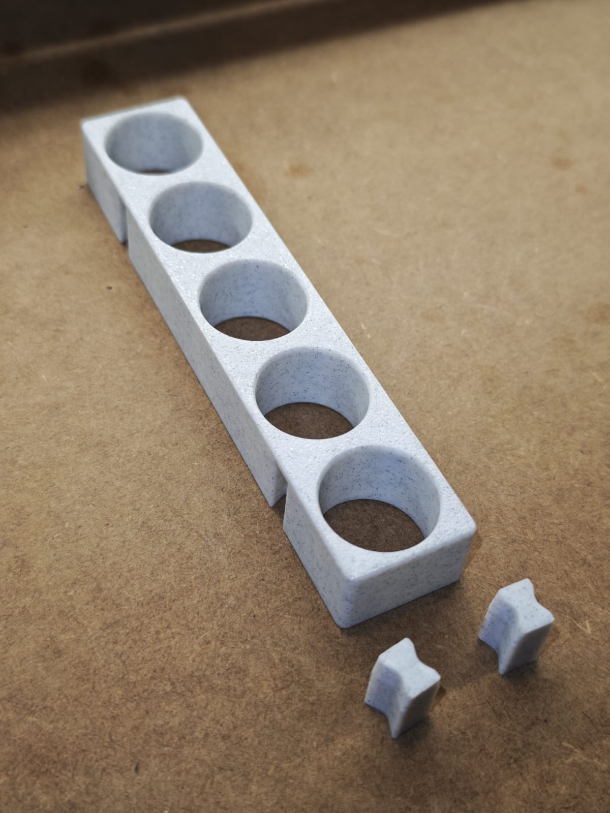 Eine hellgraue New Plastics Steinchen-Garage Modulplatte 1-3 x5 für 5 bis 15 Farbdosen Rund mit vier kreisrunden Löchern liegt auf einer braunen Fläche, neben zwei kleinen, sternförmigen Teilen aus dem gleichen Material aus dem modularen Halter-System.