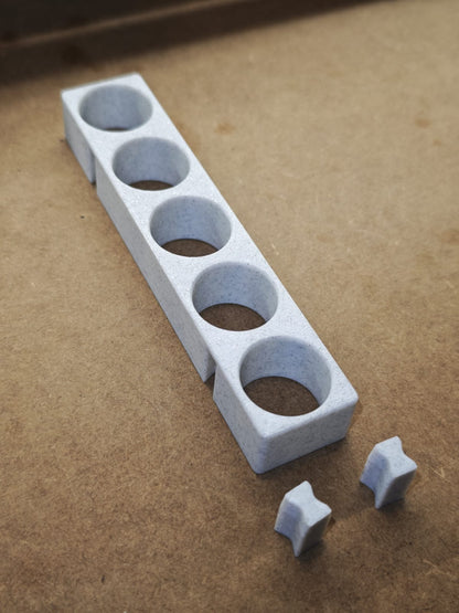 Eine hellgraue New Plastics Steinchen-Garage Modulplatte 1-3 x5 für 5 bis 15 Farbdosen Rund mit vier kreisrunden Löchern liegt auf einer braunen Fläche, neben zwei kleinen, sternförmigen Teilen aus dem gleichen Material aus dem modularen Halter-System.