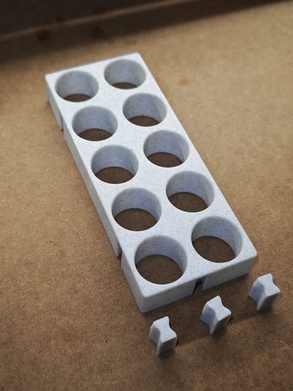 Eine weiße Steinchen-Garage Modulplatte 1-3 x5 für 5 bis 15 Farbdosen Rund von New Plastics mit acht runden Löchern liegt auf einer braunen Fläche. Davor liegen drei kreuzförmige Teile eines modularen Diamond Painting-Organisationssystems.