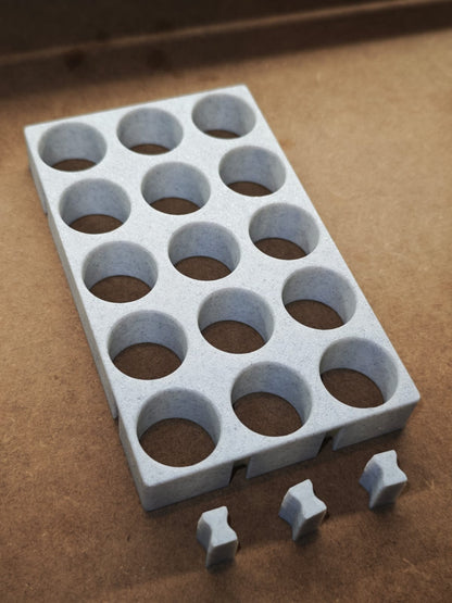 Eine graue Steinchen-Garage Modulplatte 1-3 x5 für 5 bis 15 Farbdosen Rund von New Plastics mit zwölf runden Löchern liegt auf einer braunen Fläche. Drei passende graue Dosen-Rundhalter sind vor dem Diamond Painting-Organisationssystem platziert.