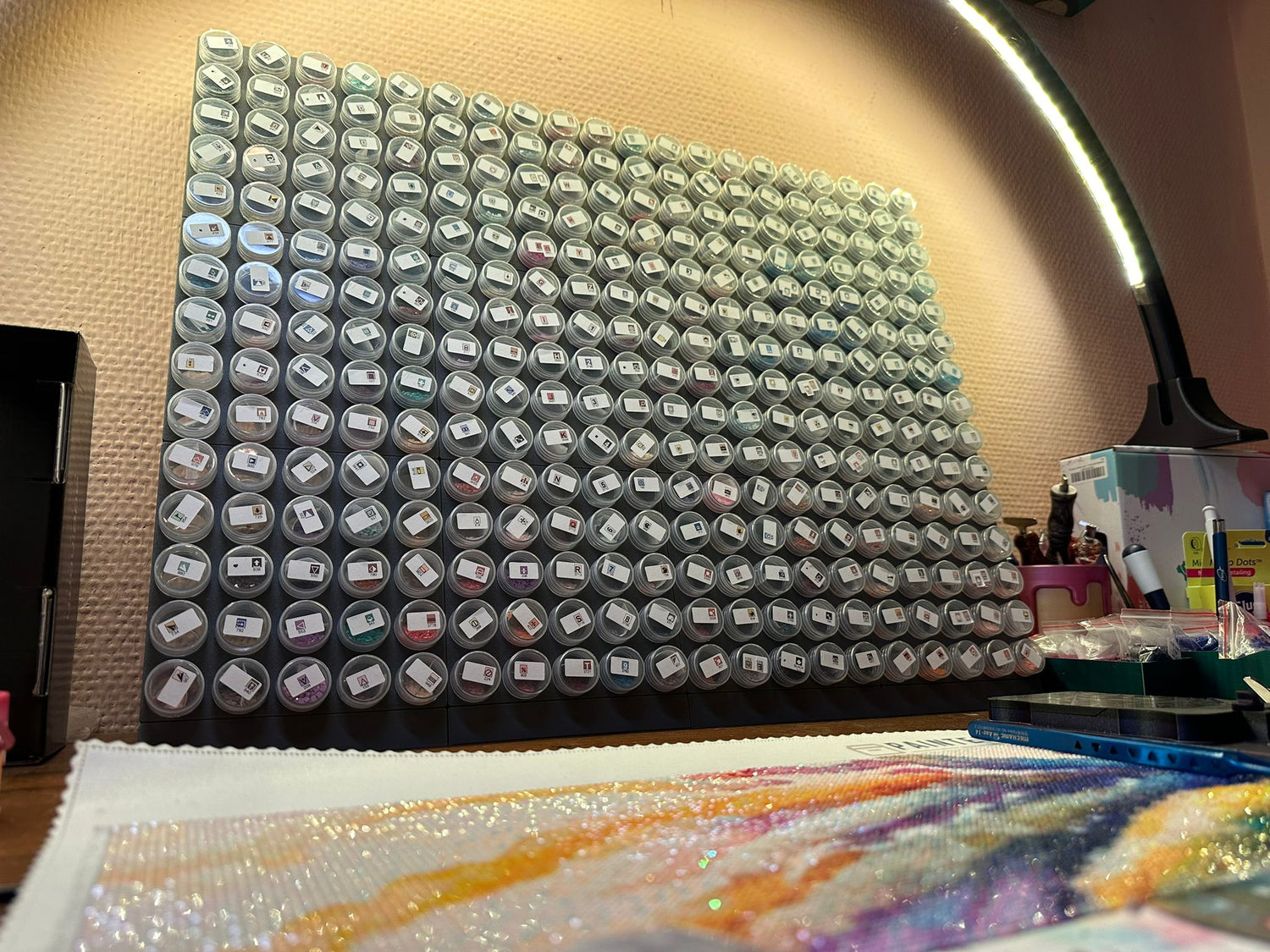 Eine Steinchen-Garage Modulplatte für 25 Farbdosen Rund von New Plastics zeigt zahlreiche kleine, beschriftete Behälter in einem Raster. Darunter ist ein farbenfrohes, halbfertiges Diamantgemälde mit Perlen und Zubehör zu sehen. Eine Deckenlampe beleuchtet den künstlerischen Arbeitsbereich.