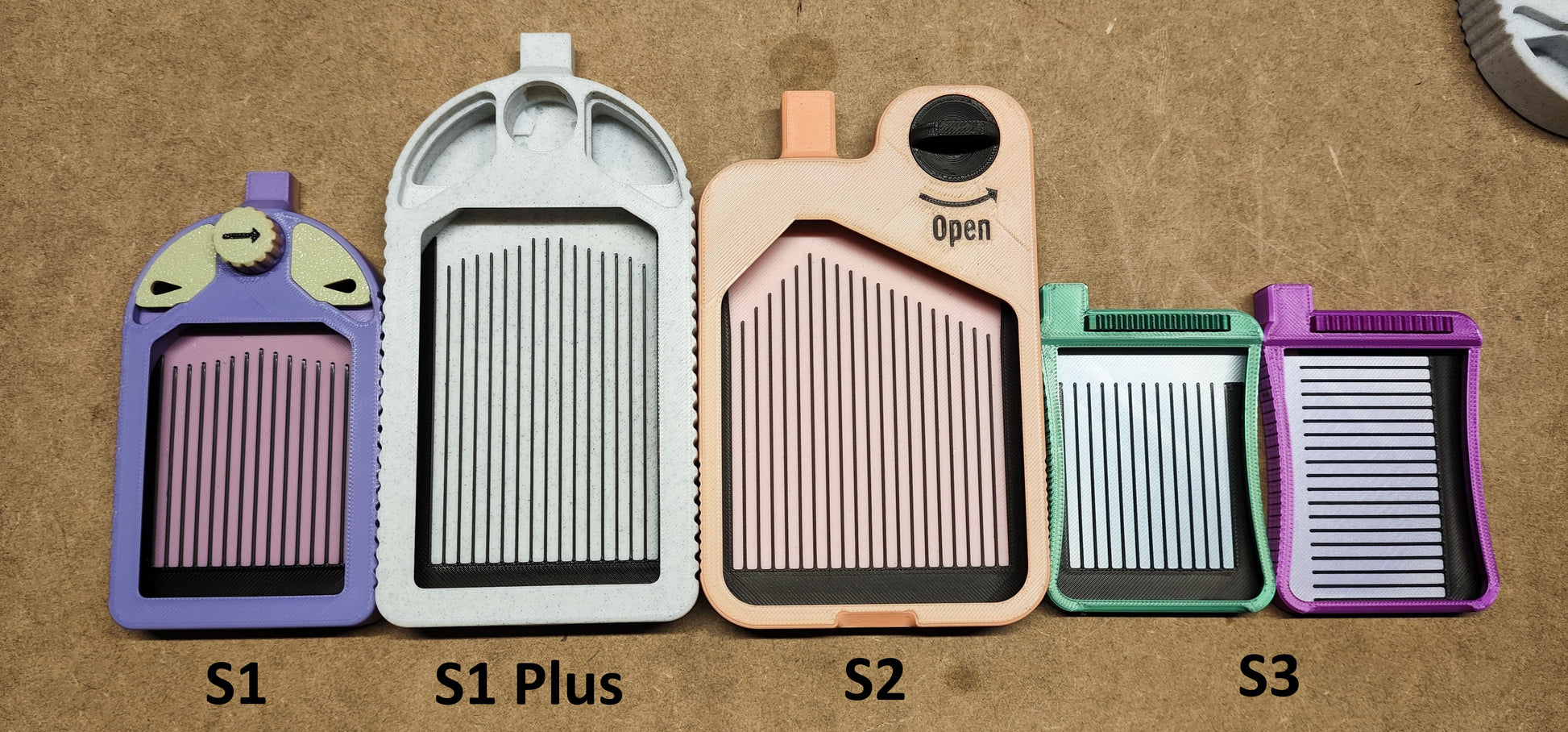 Fünf pastellfarbene Hobel von New Plastics, ideal zum Basteln oder für Diamantmalerei, werden auf Holz präsentiert. Von links nach rechts beschriftet: S1 (lila), S1 PLUS (grau), S2 (pfirsichfarben), S2 (blaugrün) und S3 (grün). Jeder Hobel hat ein Gittermuster und einen kleinen Griff.