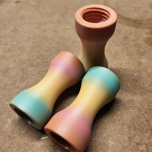 Auf einer braunen Fläche sind drei mehrfarbige, sanduhrförmige Mundstücke aus New Plastics angeordnet. Diese ergonomischen Teile zeichnen sich durch einen sanften rosa, gelben und blauen Farbverlauf mit sichtbarem Gewinde an einem Ende aus. Produkt: Schmales Griffstück für Modularen Diamond Painting Stift *Abverkauf Nur solange der Vorrat reicht*.