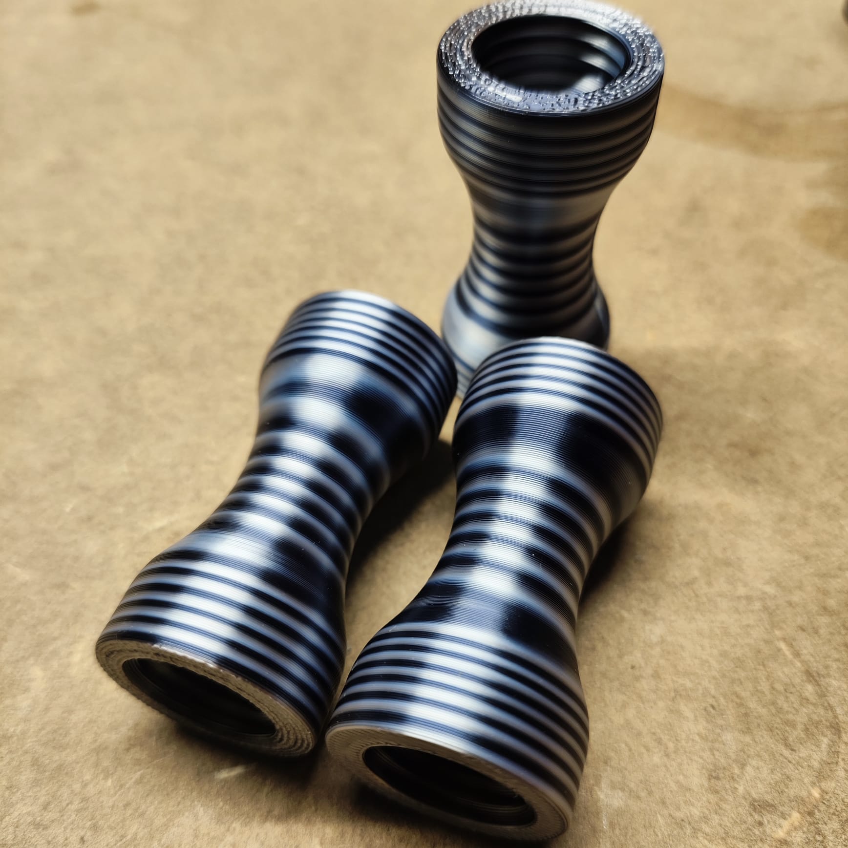 Drei metallisch gestreifte Objekte von New Plastics, die mit ihrer ergonomischen Form und geriffelten Textur an Sanduhren erinnern, liegen auf einer Kartonoberfläche. Zwei liegen, eines steht aufrecht. Alle präsentieren das *Schmales Griffstück für Modularen Diamond Painting Stift*.