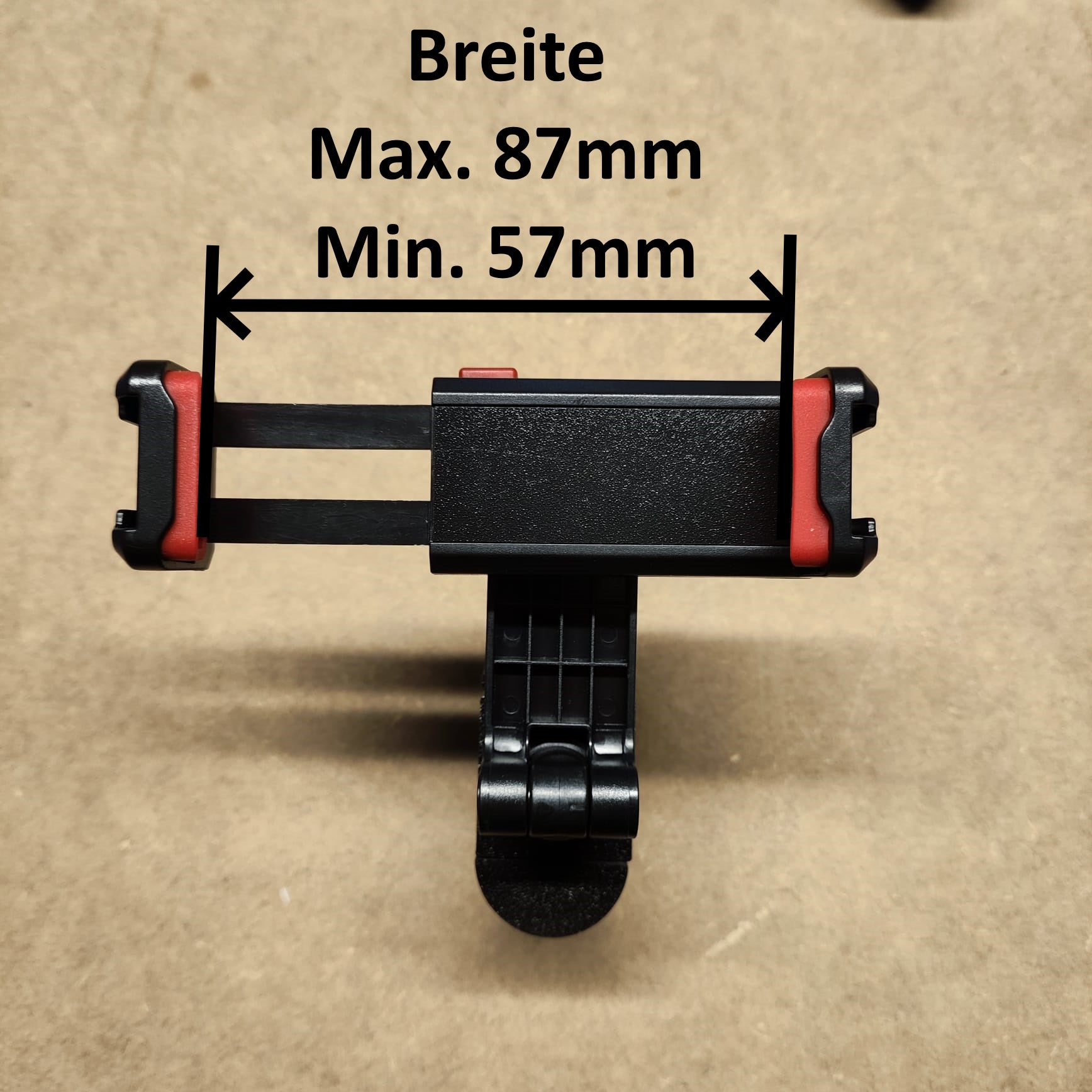 Die schwarz-rote Handyhalterung von New Plastics, entworfen für die Original Steinchen Garage, ist auf braunem Hintergrund abgebildet. Ihre einstellbare Breite („Breite Max. 87mm, Min. 57mm“) wird in ausgefahrener Position hervorgehoben und zeigt ihre perfekte Passform für die sichere Befestigung von Geräten.