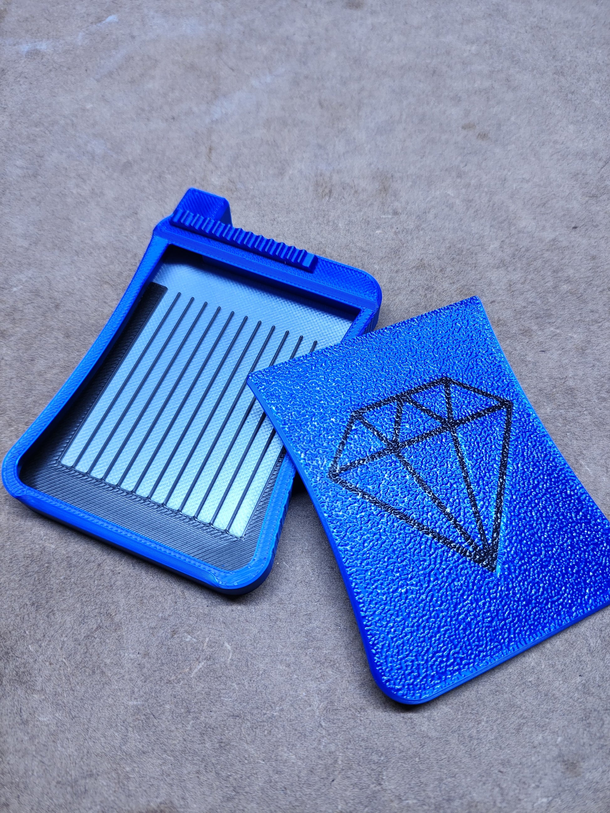 Das New Plastics Diamond Painting Schiffchen S3 verfügt über ein blaues Kunststoffgehäuse mit einem kammartigen Einsatz auf der linken Seite und einer strukturierten Abdeckung mit einer Diamantillustration auf der rechten Seite. Sein Präzisionsdesign im FDM-Verfahren wird durch einen neutralen Stoffhintergrund hervorgehoben und eignet sich ideal zur Aufbewahrung von Bastelarbeiten.