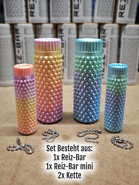 Vier bunte, strukturierte zylindrische Behälter und Ketten mit der Aufschrift „Set besteht aus: 1x Reiz-Bar, 1x Reiz-Bar mini, 2x Kette“ zur taktilen Stimulation durch das Reiz-Bar-Set von New Plastics liegen auf einer Oberfläche. Weitere ähnliche Gegenstände erscheinen im unscharfen Hintergrund.