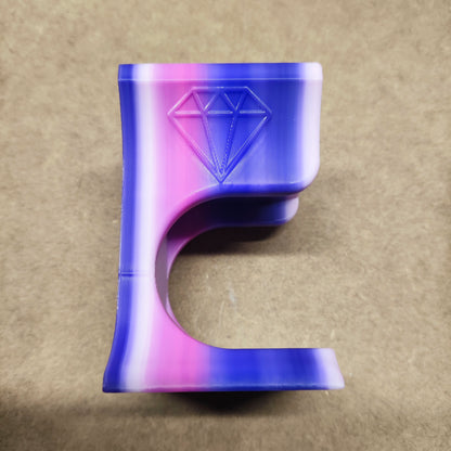 Ein 3D-gedrucktes Objekt mit einem violetten, rosa und weißen Farbverlauf weist ein geprägtes Rautensymbol auf. Seine einzigartige geschwungene Form ähnelt dem Diamond Painting Trichter für runde Dosen von New Plastics und ist auf einem schlichten beigen Hintergrund fotografiert.