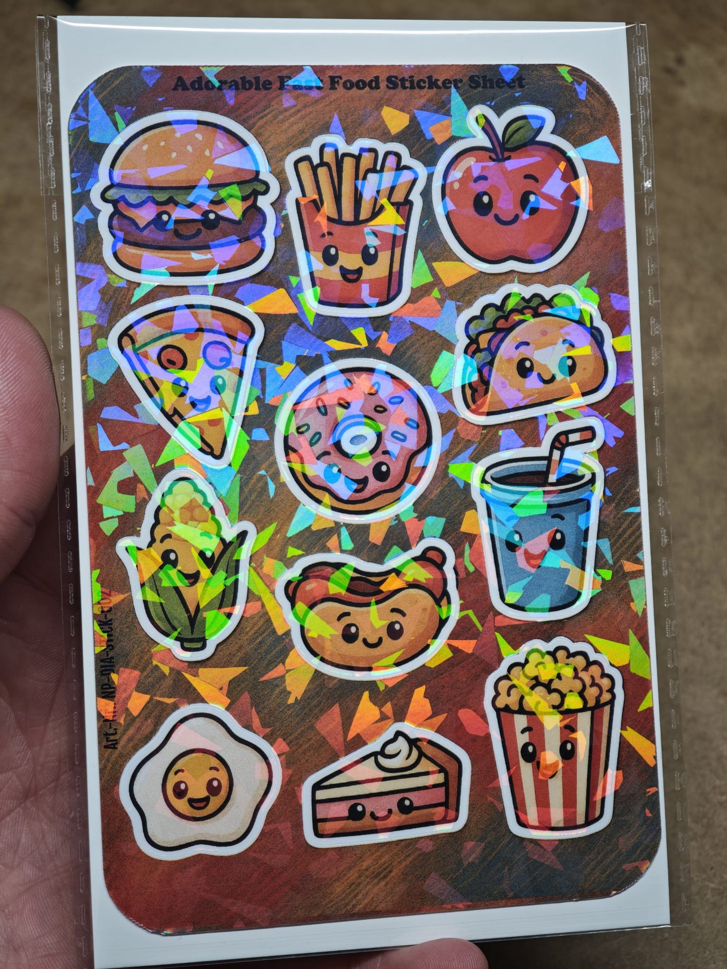 Eine Hand hält den „Adorable Fast Food Sticker Sheet mit 12 Stickern ca. DIN A6“ von New Plastics, mit süßen holografischen Burgern, Pommes, Pizza, Donuts und mehr – perfekt für DIY-Projekte oder Ihre Sticker Sheets-Sammlung.