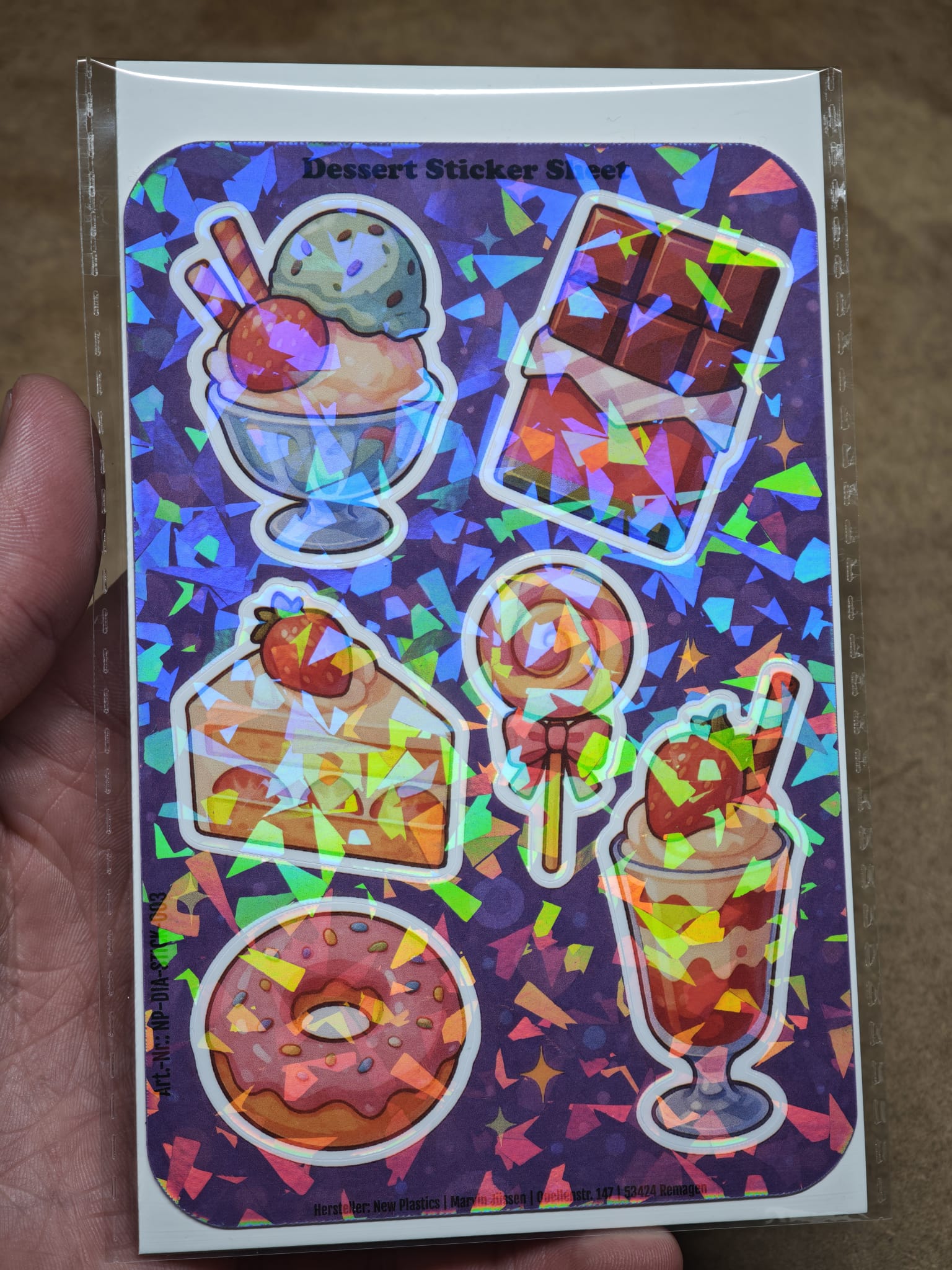 Eine Hand hält den New Plastics Dessert Sticker Sheet mit 6 Stickern ca. DIN A6 – mit Eis, Schokolade, Kuchen, Lutscher, Donut und Parfait – für DIY-Projekte vor einem leuchtend violetten Hintergrund.
