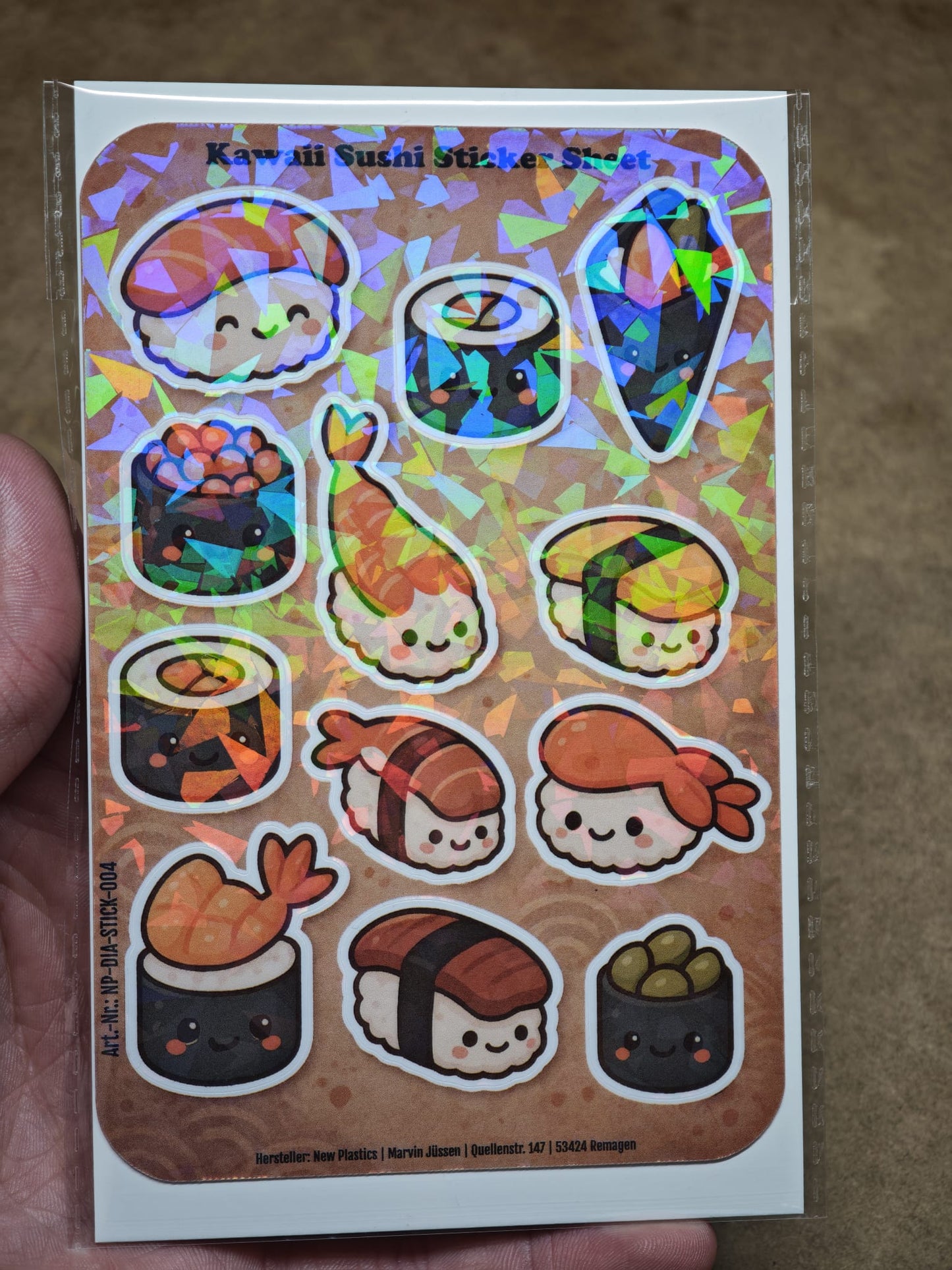 Eine Hand hält den Kawaii Sushi Sticker Sheet mit 12 Stickern ca. DIN A6 von New Plastics, mit süßen Nigiri-, Maki- und Onigiri-Aufklebern – ideal für DIY-Projekte – vor einem braun gemusterten Hintergrund.