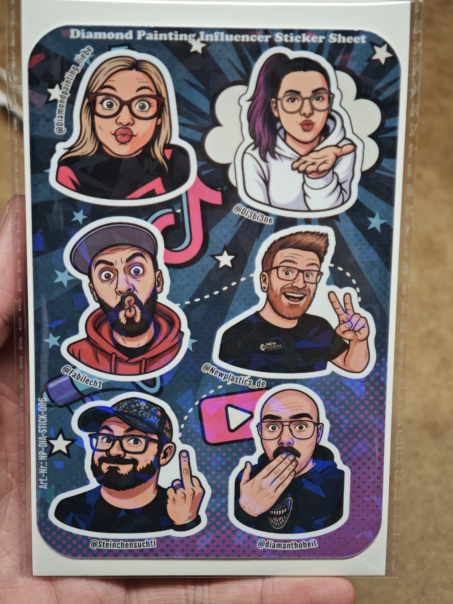 Eine Hand zeigt den New Plastics Diamond Painting „Influencer“ Stickerbogen (ca. DIN A6) mit 6 ausdrucksstarken Charakter-Stickern auf blauem Starburst-Hintergrund – ideal für DIY-Projekte, jeweils inklusive Social-Media-Handle.