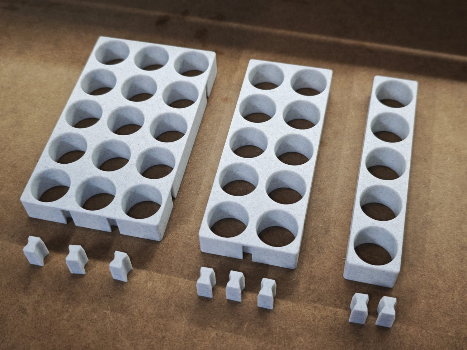 Drei graue Steinchen-Garage Modulplatten 1-3 x5 für 5 bis 15 Farbdosen Rund von New Plastics, jeweils mit kreisrunden Löchern und paarweise passenden Teilen, sind auf einer braunen Oberfläche angeordnet, ideal für Diamond Painting-Organisationssysteme.