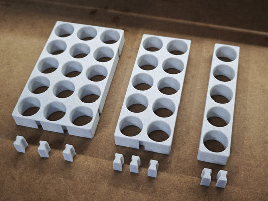 Drei graue Steinchen-Garage Modulplatten 1-3 x5 für 5 bis 15 Farbdosen Rund von New Plastics, jeweils mit kreisrunden Löchern und paarweise passenden Teilen, sind auf einer braunen Oberfläche angeordnet, ideal für Diamond Painting-Organisationssysteme.
